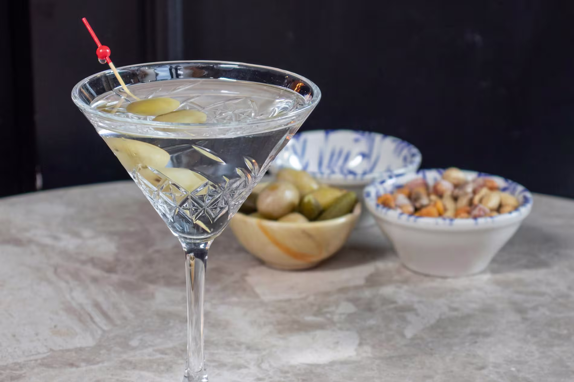 A martini garnished with olives sits on a table beside small bowls of olives, pickles, and mixed nuts.