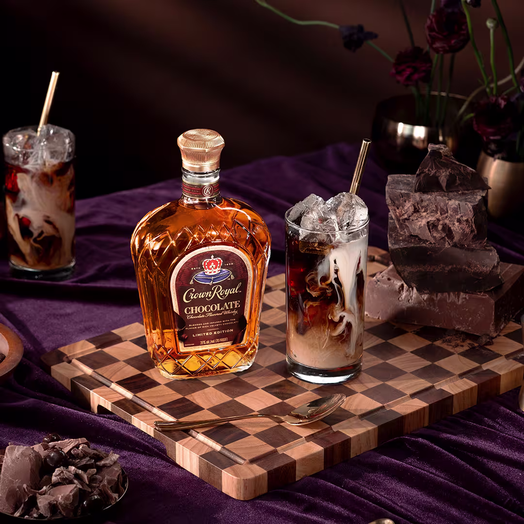 A bottle of Crown Royal Chocolate whisky, a glass of iced mixed drink, and chunks of chocolate are arranged on a checkered wooden board with a purple tablecloth.