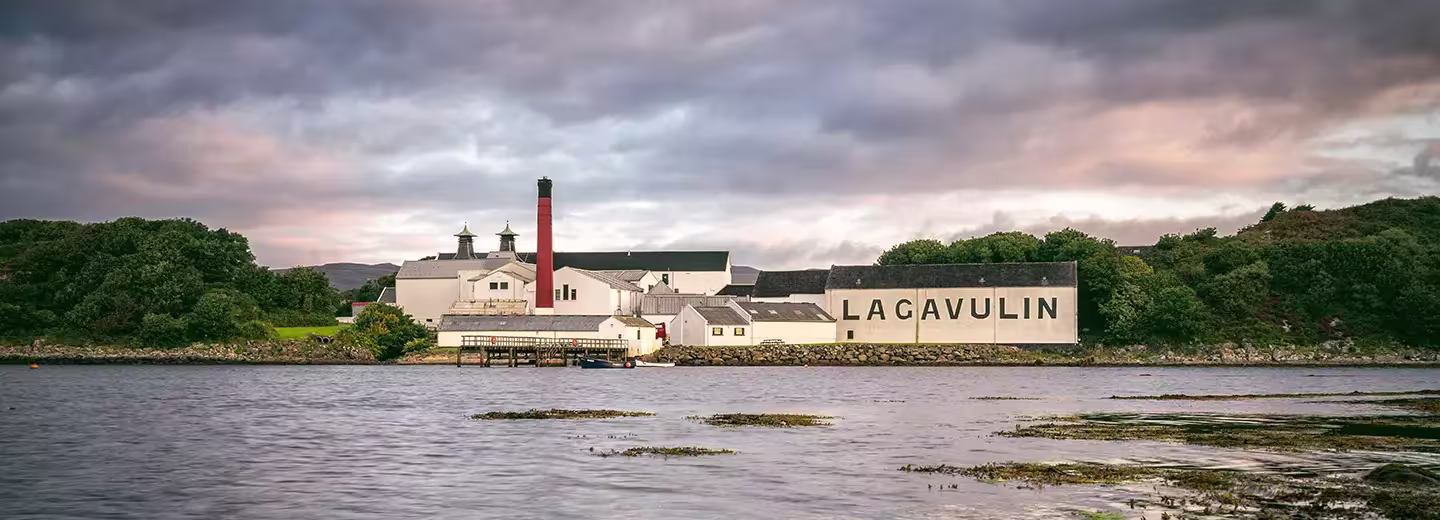 A distillery complex labeled Lagavulin sits by the water’s edge with trees in the background under a cloudy sky.