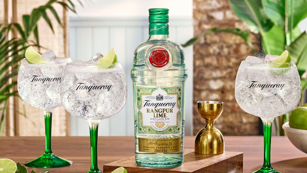A bottle of Tanqueray Rangpur Lime gin is displayed with three gin and tonic glasses, a gold jigger, and lime wedges on a wooden tray.