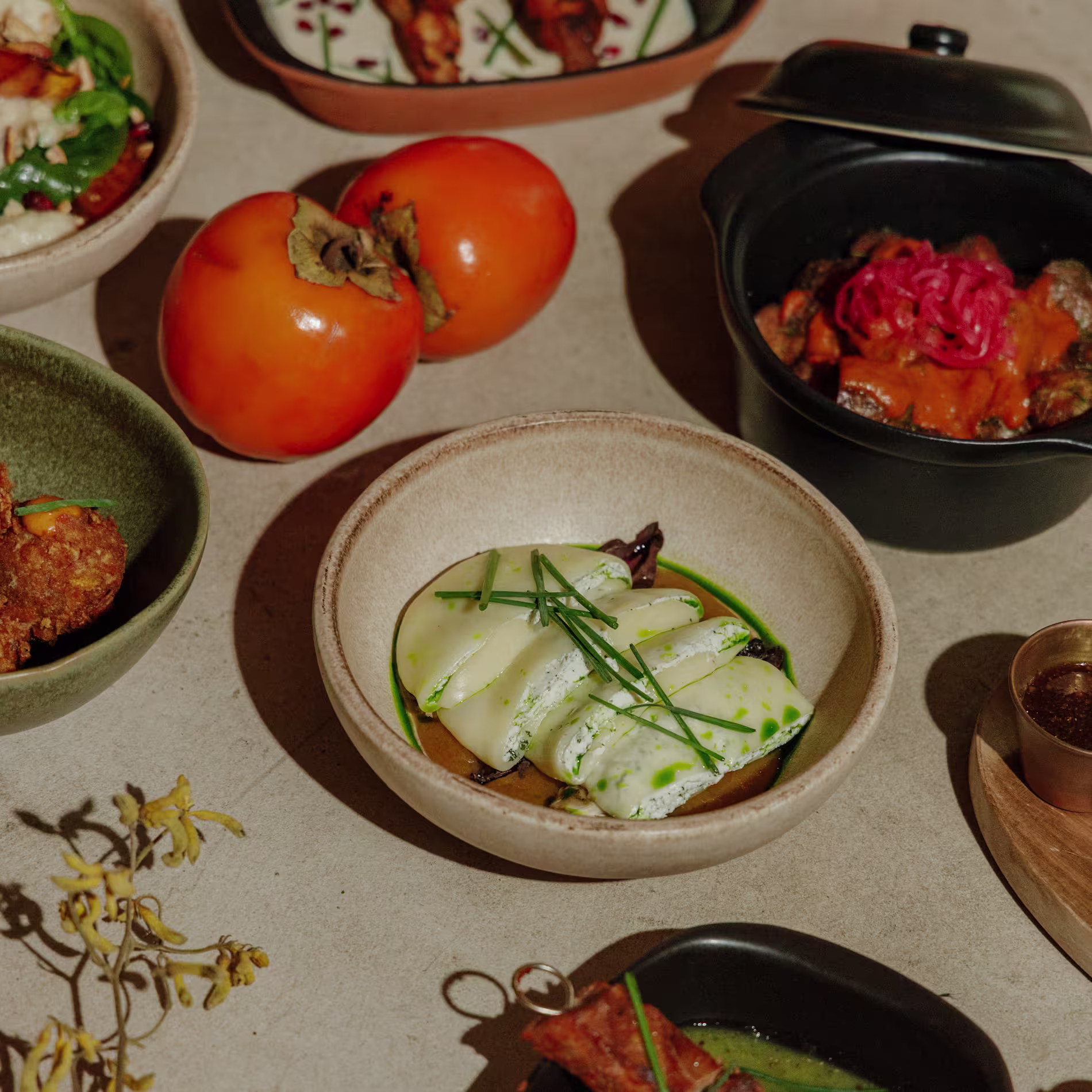 La foto muestra una variedad de Persimmons. Hay varios platos con diferentes presentaciones de alimentos, incluyendo vegetales en rodajas en un plato y carne a la parrilla en otro.