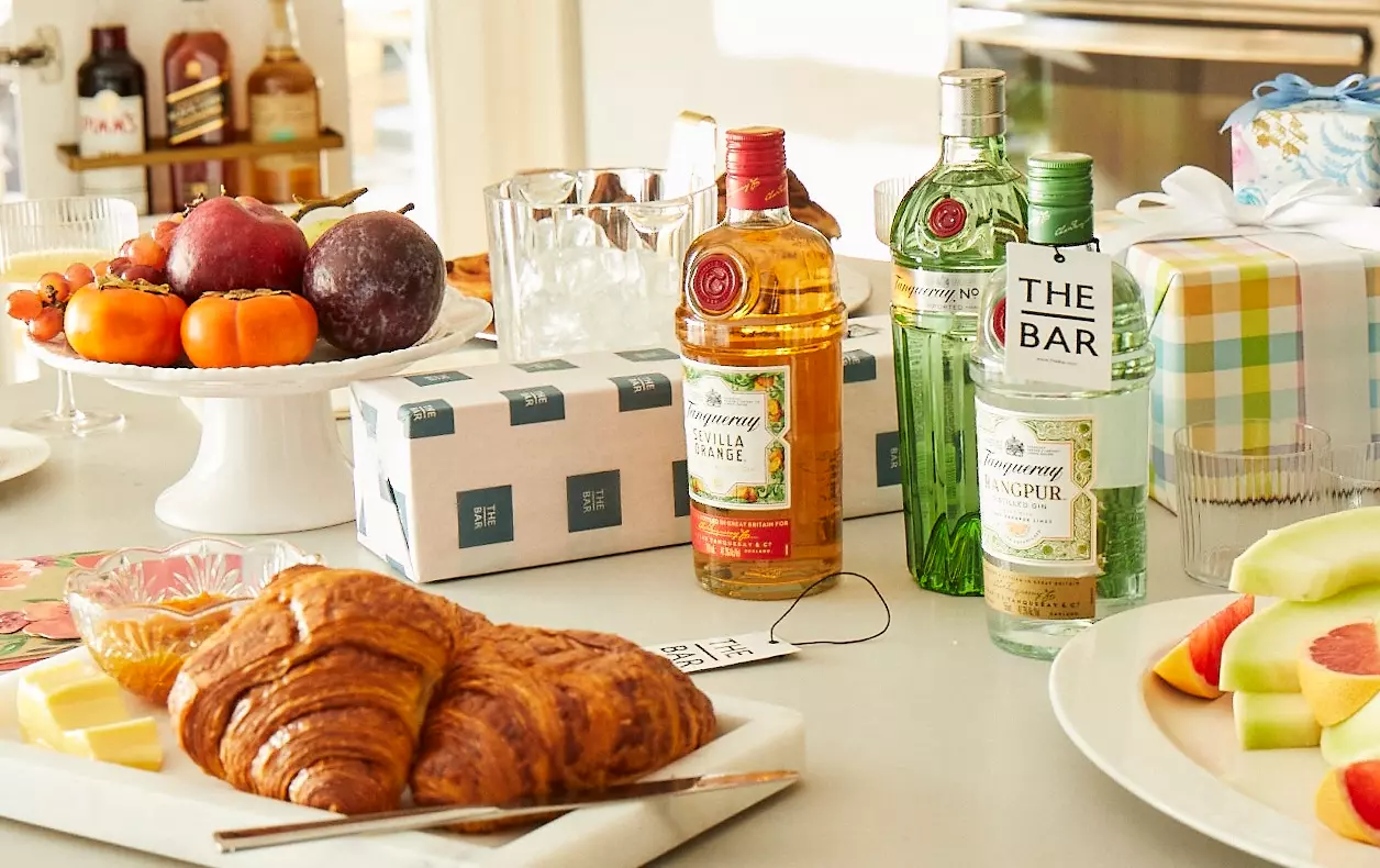 A breakfast table with croissants, fruit, cheese, wrapped gifts, and three bottles of Tanqueray gin arranged near glasses and ice.
