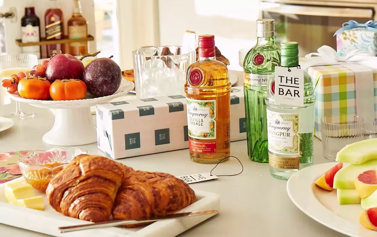 A breakfast table with croissants, fruit, cheese, wrapped gifts, and three bottles of Tanqueray gin arranged near glasses and ice.