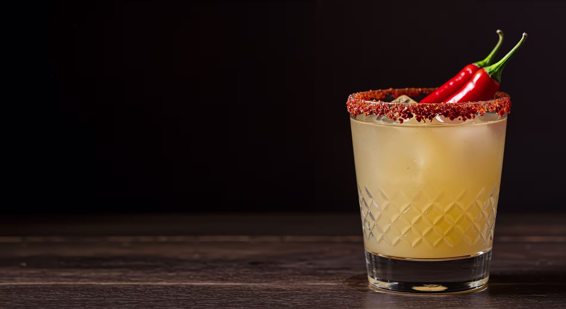 A cocktail in a glass with a chili powder rim, ice, and two red chili peppers as garnish, set against a dark background.