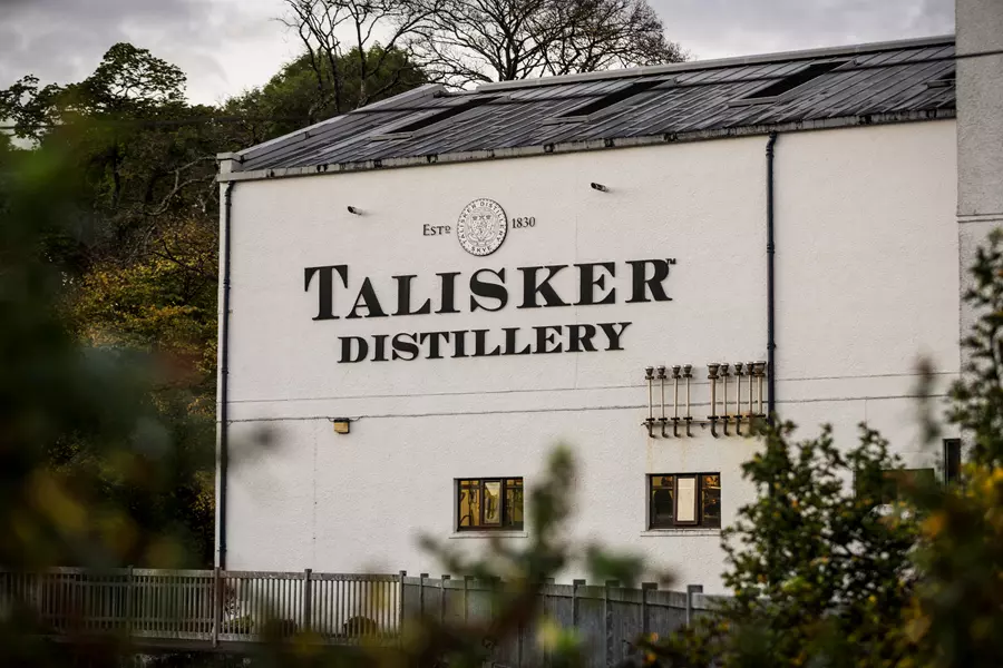 Exterior view of the Talisker Distillery building with large signage and a few windows, surrounded by greenery.