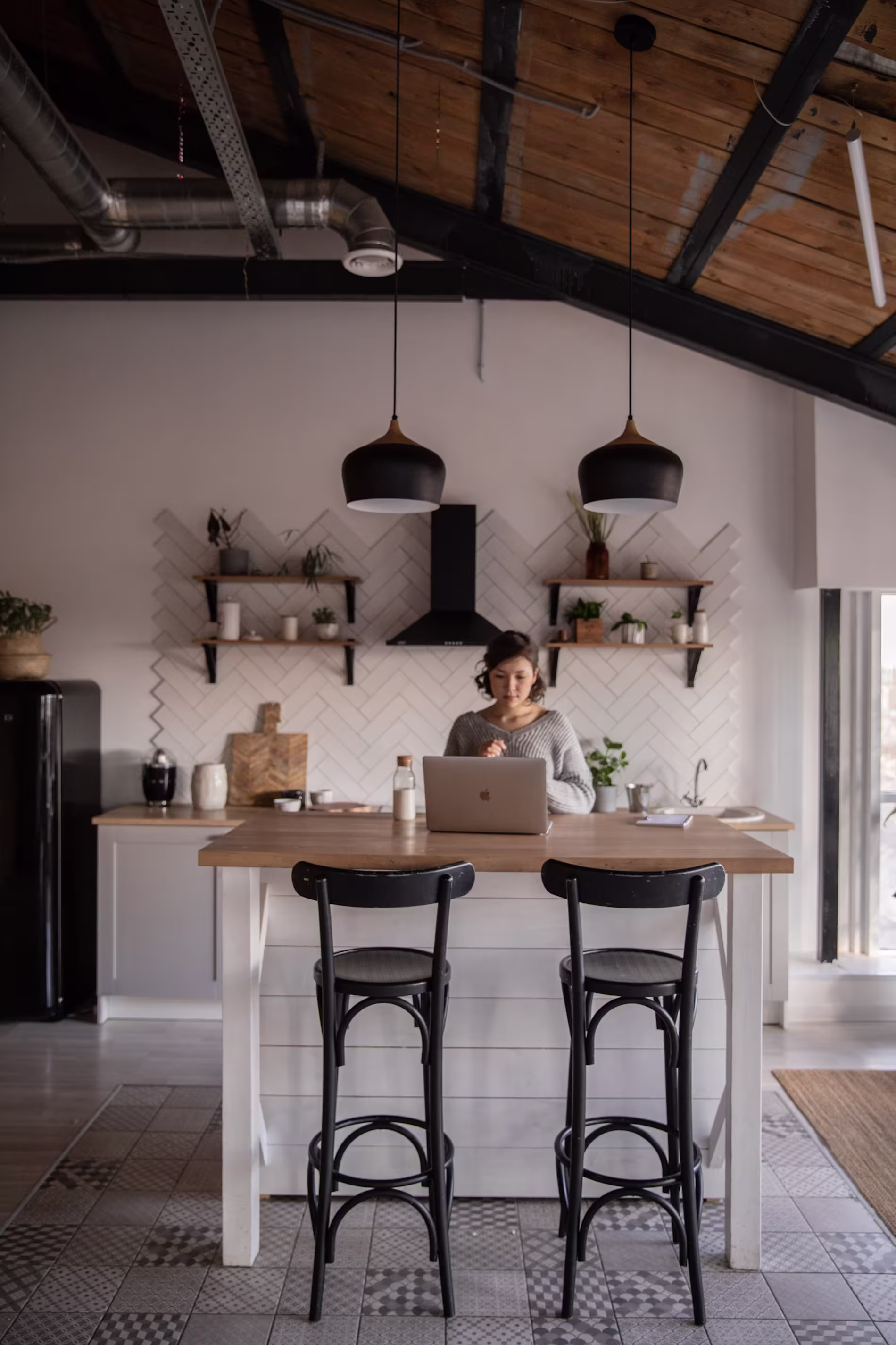 A person works on a laptop at a kitchen island in a modern kitchen with hanging lights, open shelves, and wooden ceiling beams.