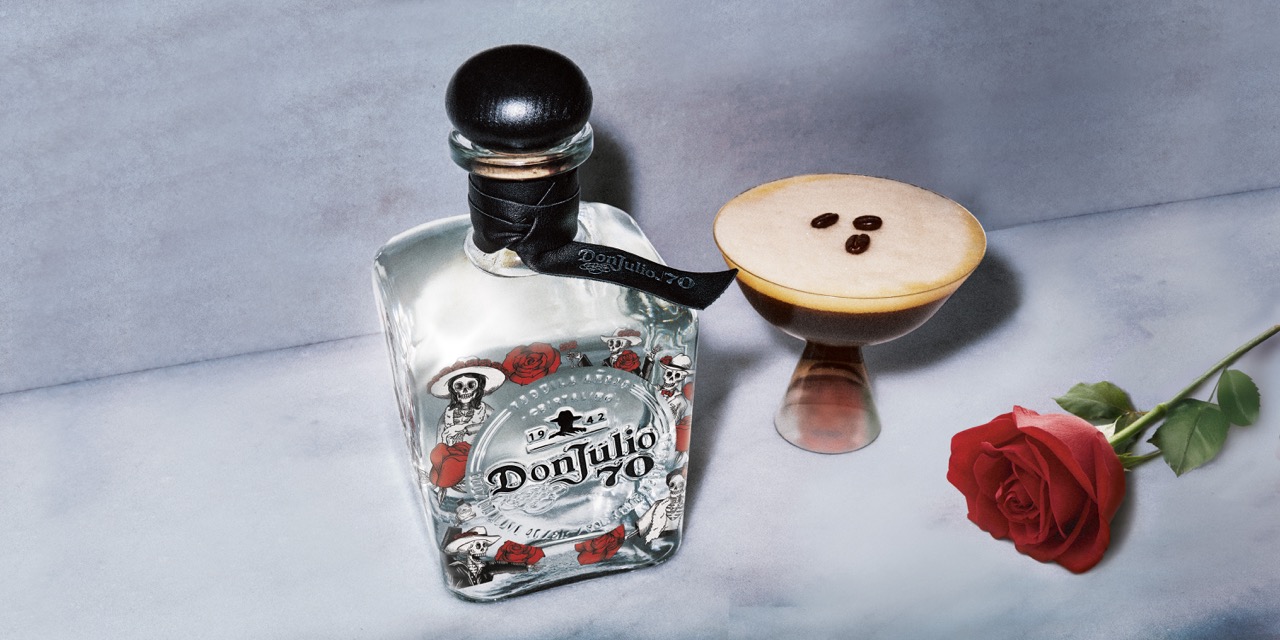 A bottle of Don Julio 70 tequila next to a cocktail garnished with three coffee beans and a red rose on a light marble surface.