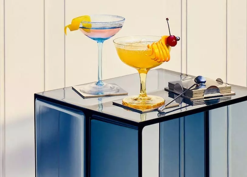 Two cocktail glasses with colorful drinks and garnishes sit on a glass table, next to a stack of napkins and a cocktail stirring spoon.