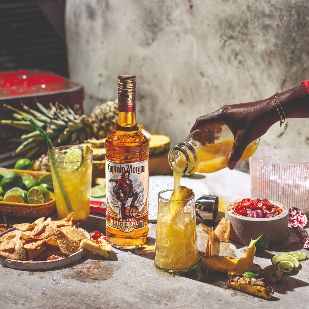 Hand pouring a Captain Morgan Spiced Rum cocktail with a bottle of Captain Morgan, tortilla chips, salsa, limes, and fresh pineapple on a table