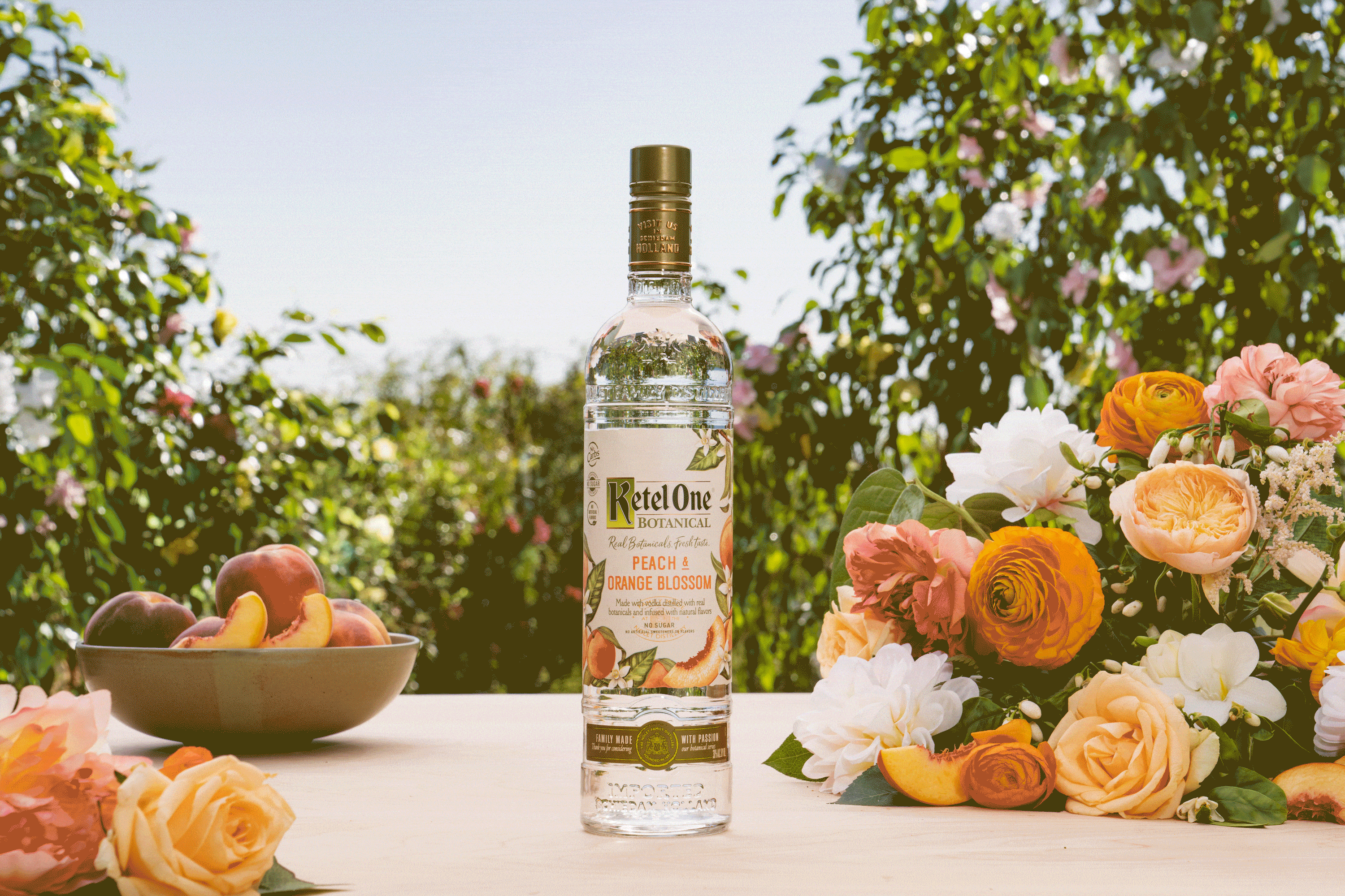 A bottle of Ketel One Botanical Peach & Orange Blossom vodka stands on a table with a bowl of peaches and orange flowers, set outdoors with greenery in the background.