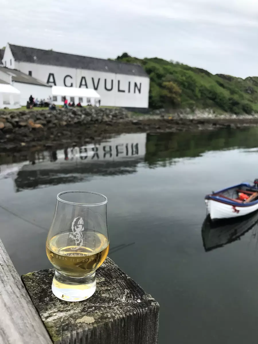 A glass of whisky is placed on a wooden railing by the water, with a boat and the Lagavulin distillery building in the background.