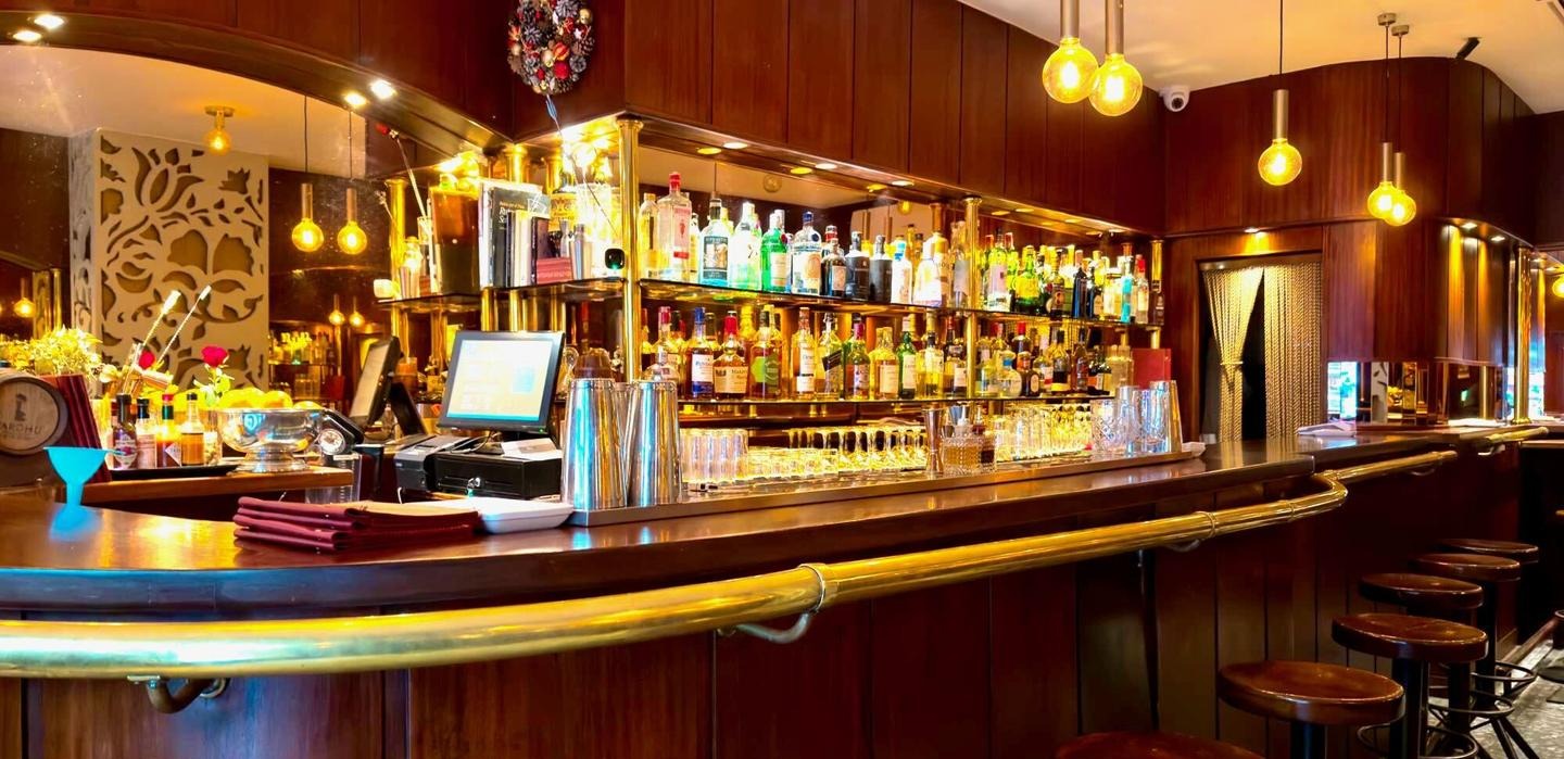 La imagen muestra una barra de bar de estilo clásico, con estanterías llenas de botellas y una iluminación cálida. El ambiente es elegante y acogedor, con taburetes de madera alrededor de la barra.
