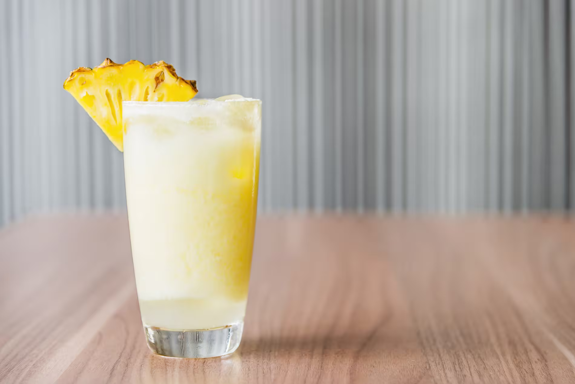 A glass of frozen yellow beverage garnished with a pineapple slice sits on a wooden table with a blurred gray background.