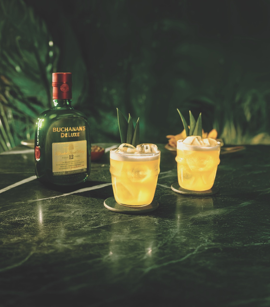 A bottle of Buchanan’s Deluxe whisky and two yellow cocktails with ice and pineapple garnish sit on a dark green marble table.