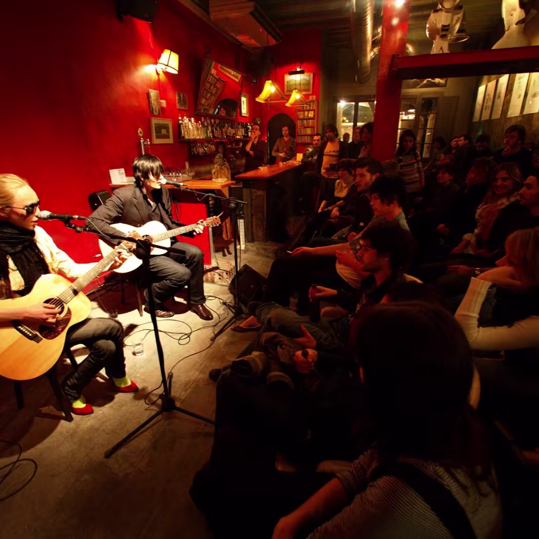 La imagen muestra a dos músicos tocando guitarras acústicas en un ambiente íntimo, rodeados de un público atento en un local con paredes rojas y una atmósfera cálida.