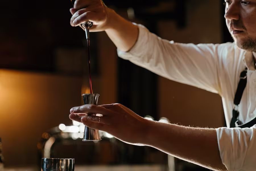 A person pours a dark liquid from a bottle into a jigger, preparing a drink at a bar.