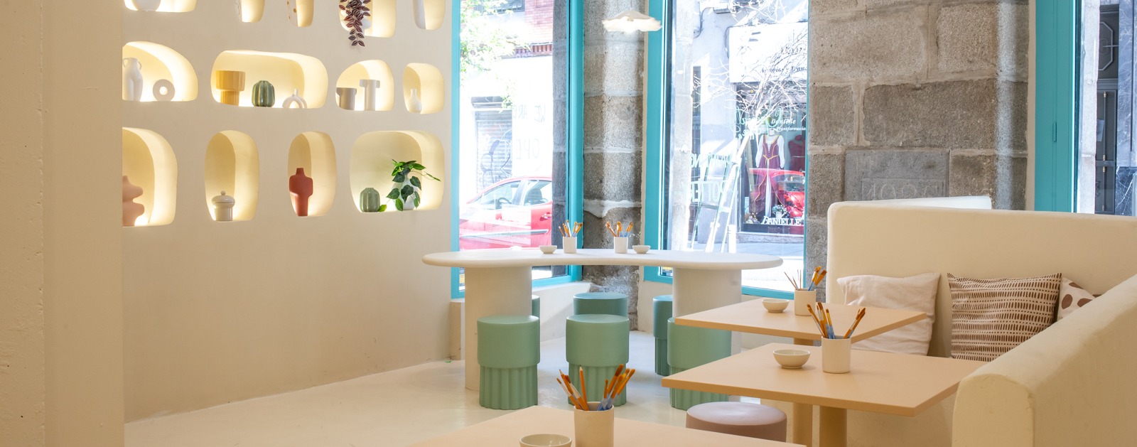 Interior de Mama Pottery Café con decoración blanca y luminosa, nichos con cerámica decorativa, ventanas azul turquesa y mesas listas para pintar.
