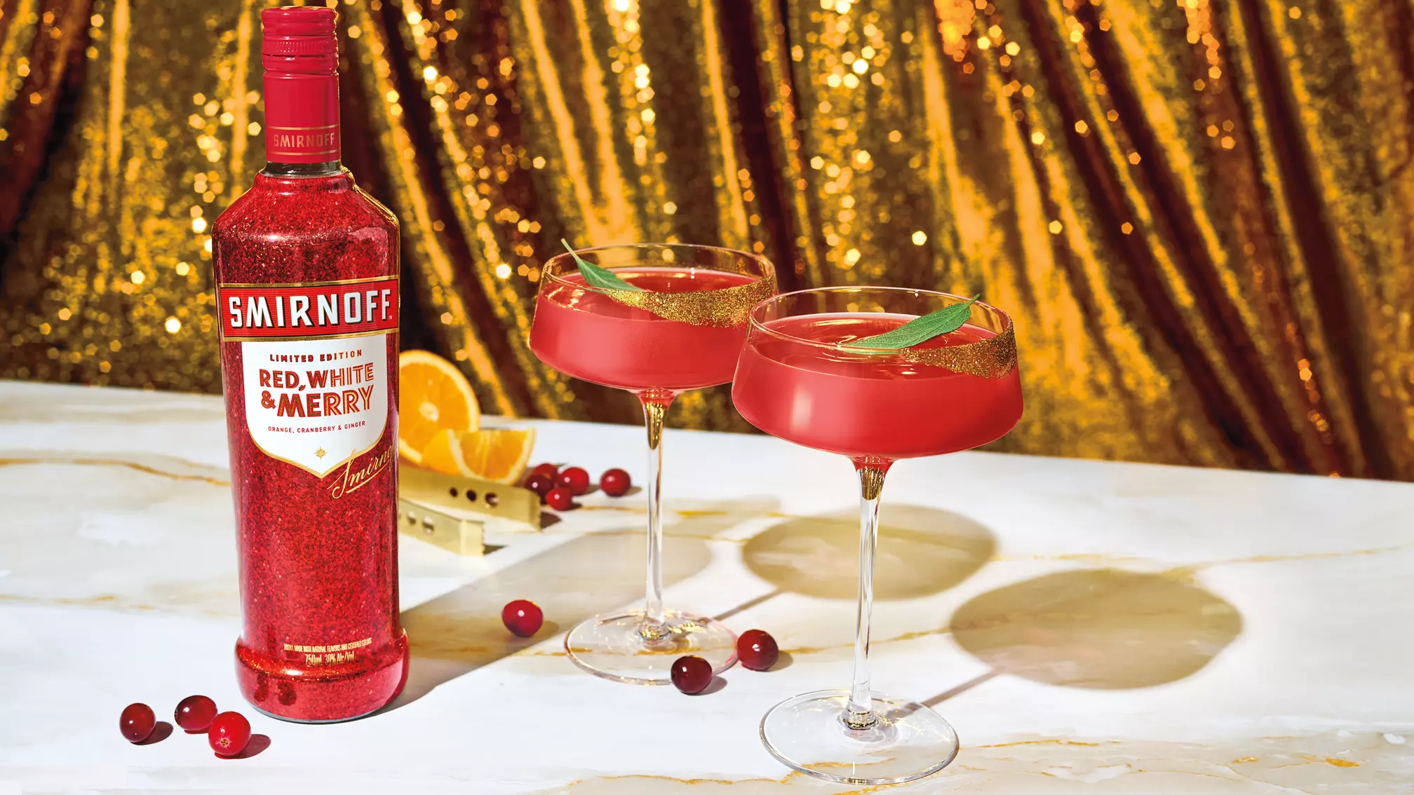 A bottle of Smirnoff Red, White & Merry vodka stands next to two red cocktails in coupe glasses, with cranberries, orange slices, and a gold sequin backdrop.