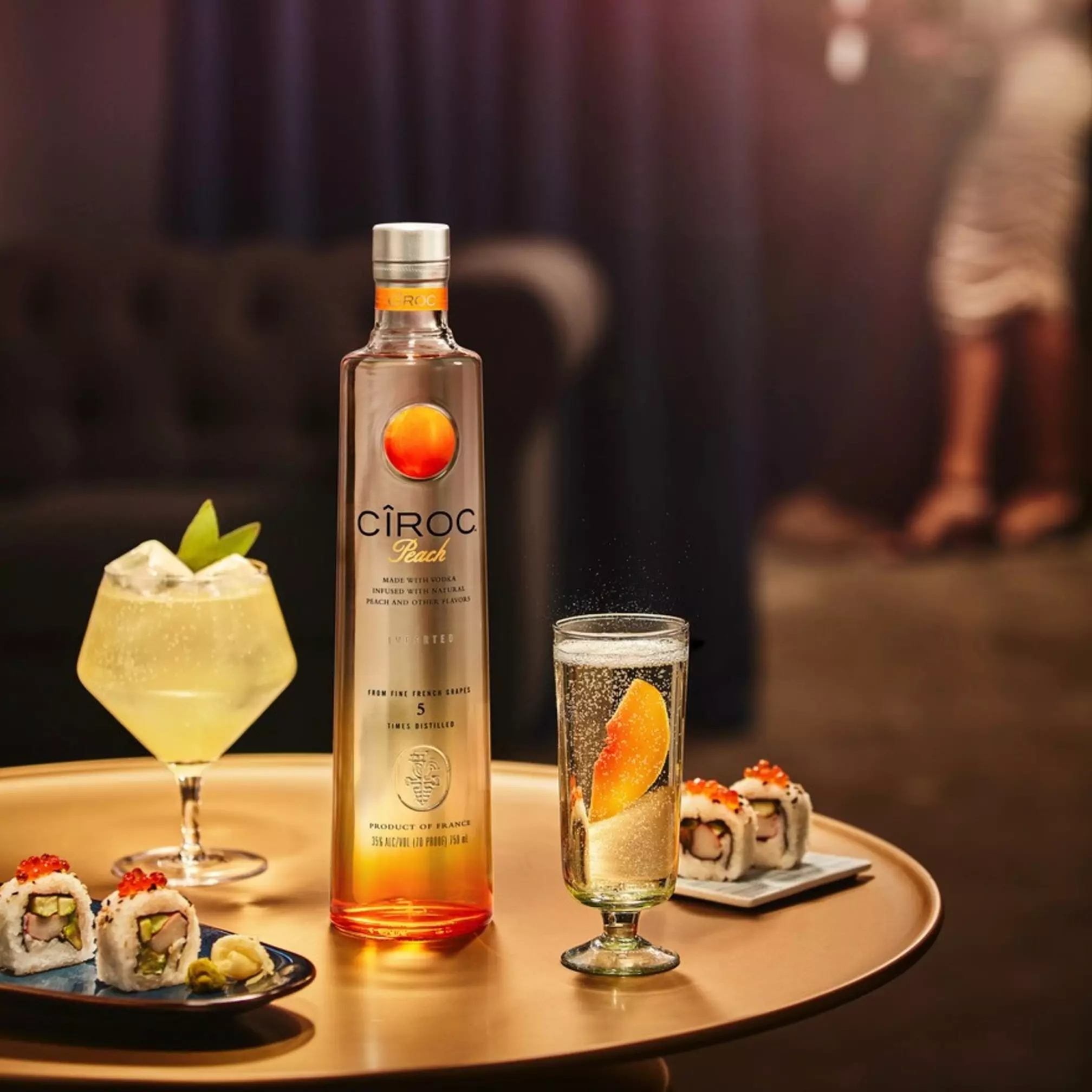 A bottle of Cîroc Peach vodka is on a table with a cocktail, a glass with an orange slice, and sushi rolls. Two blurred figures are in the background.