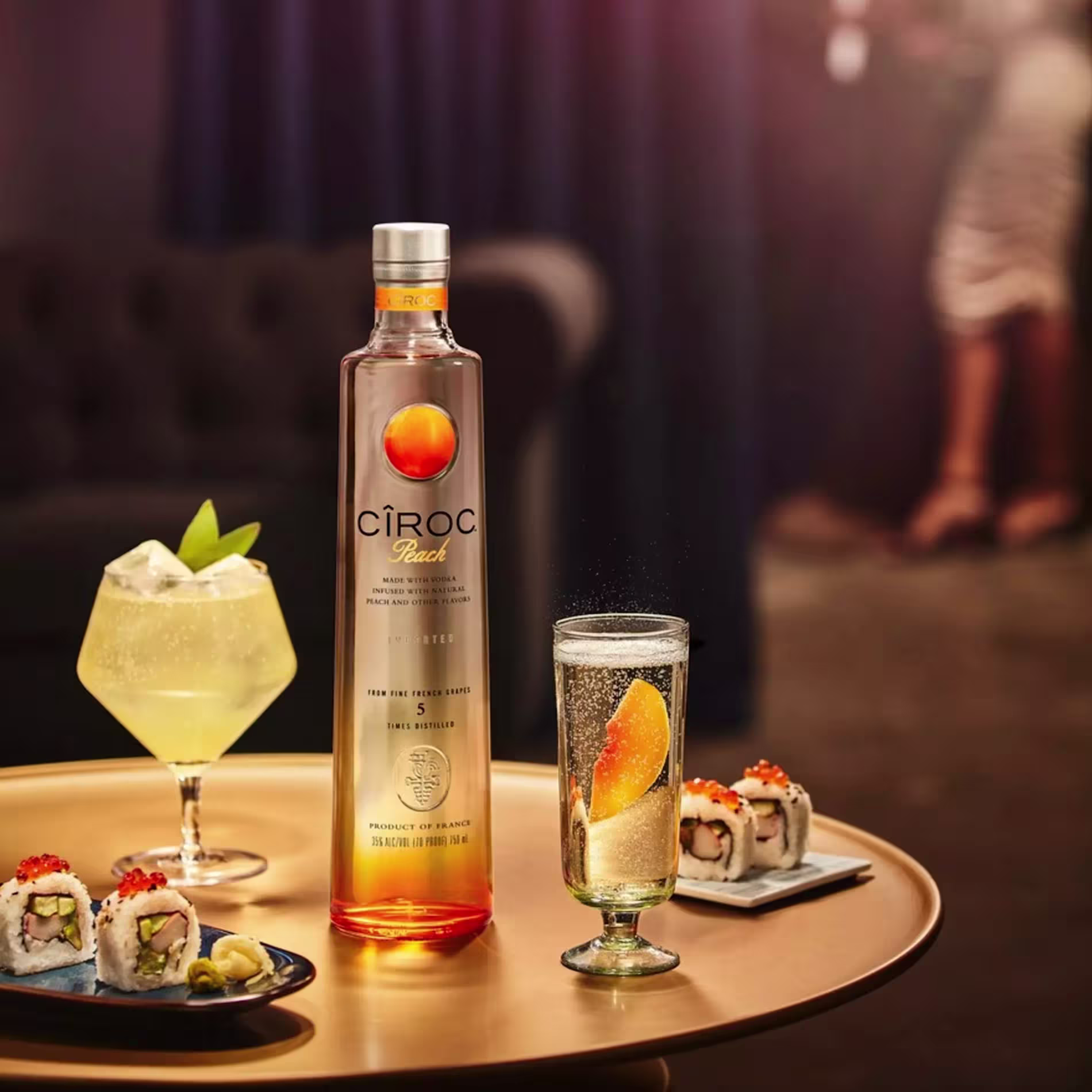 A bottle of Cîroc Peach vodka is on a table with a cocktail, a glass with an orange slice, and sushi rolls. Two blurred figures are in the background.