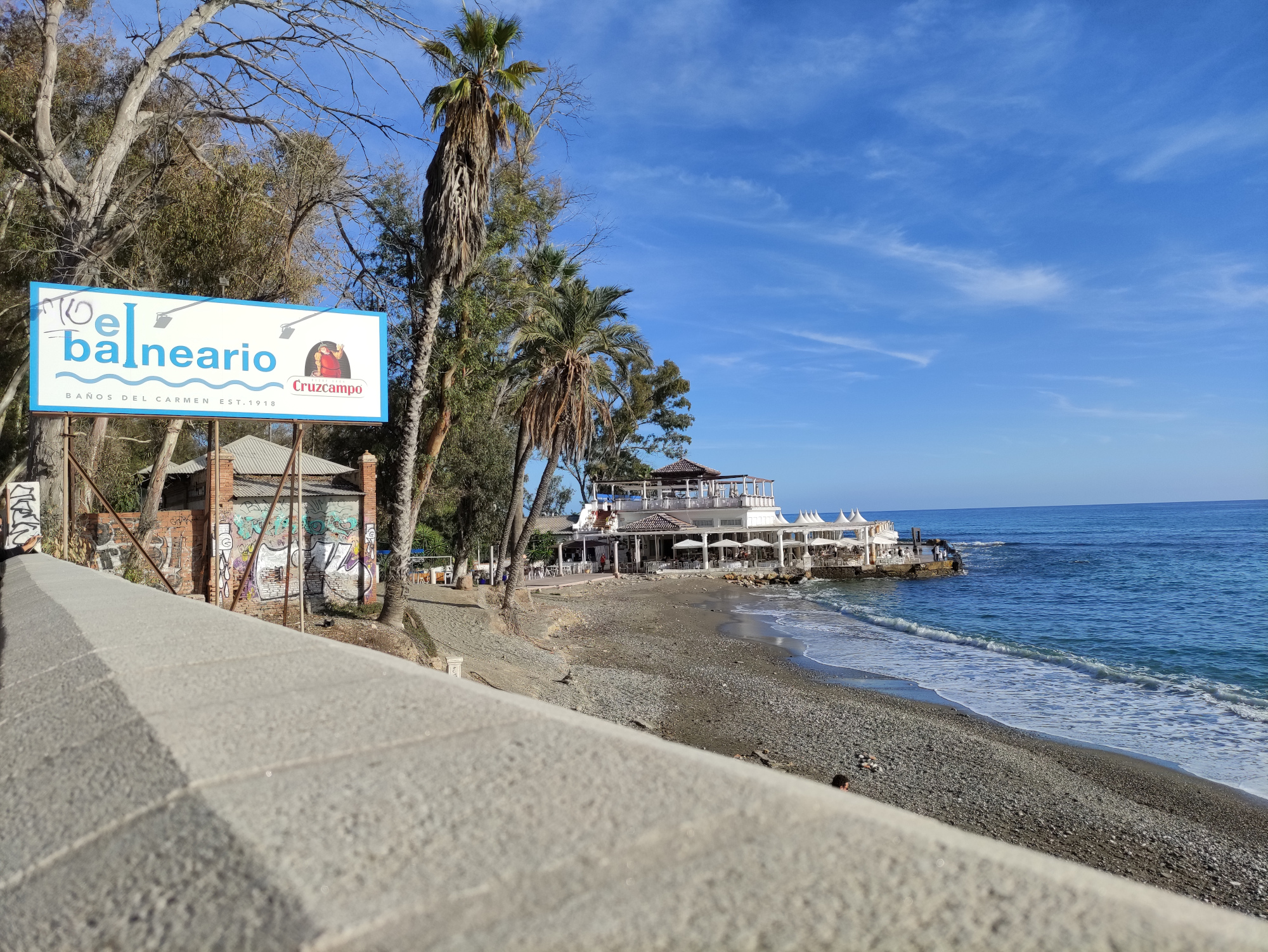 La imagen muestra una vista de una playa con un cielo azul claro y un mar tranquilo. Hay un cartel que dice “El Balneario” Se pueden ver palmeras y una estructura de edificio con un área de asientos al aire libre que se extiende sobre el agua.