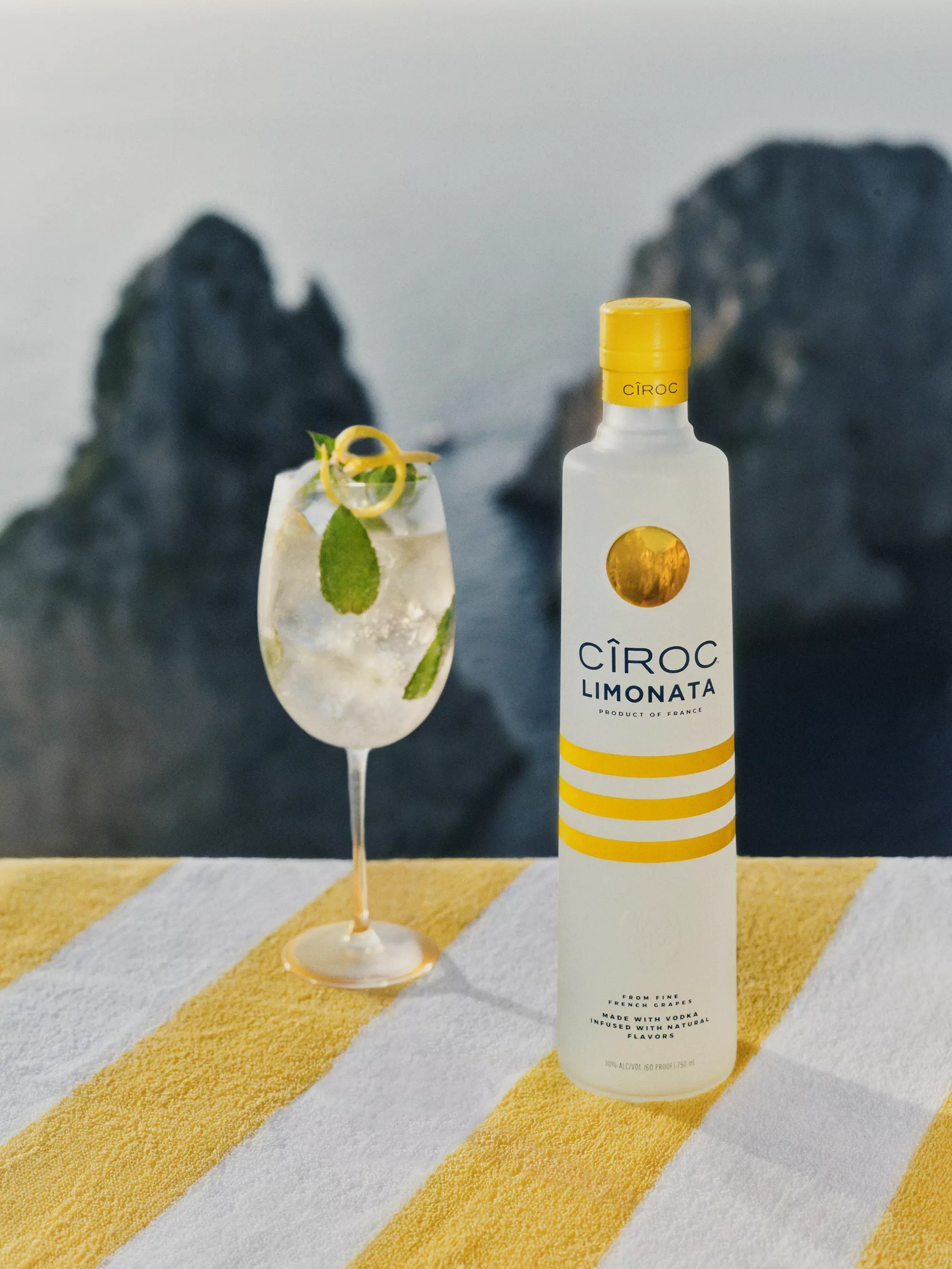 A bottle of Cîroc Limonata sits on a yellow and white striped towel next to a cocktail glass with ice, mint, and lemon, with rocky cliffs and water in the blurred background.