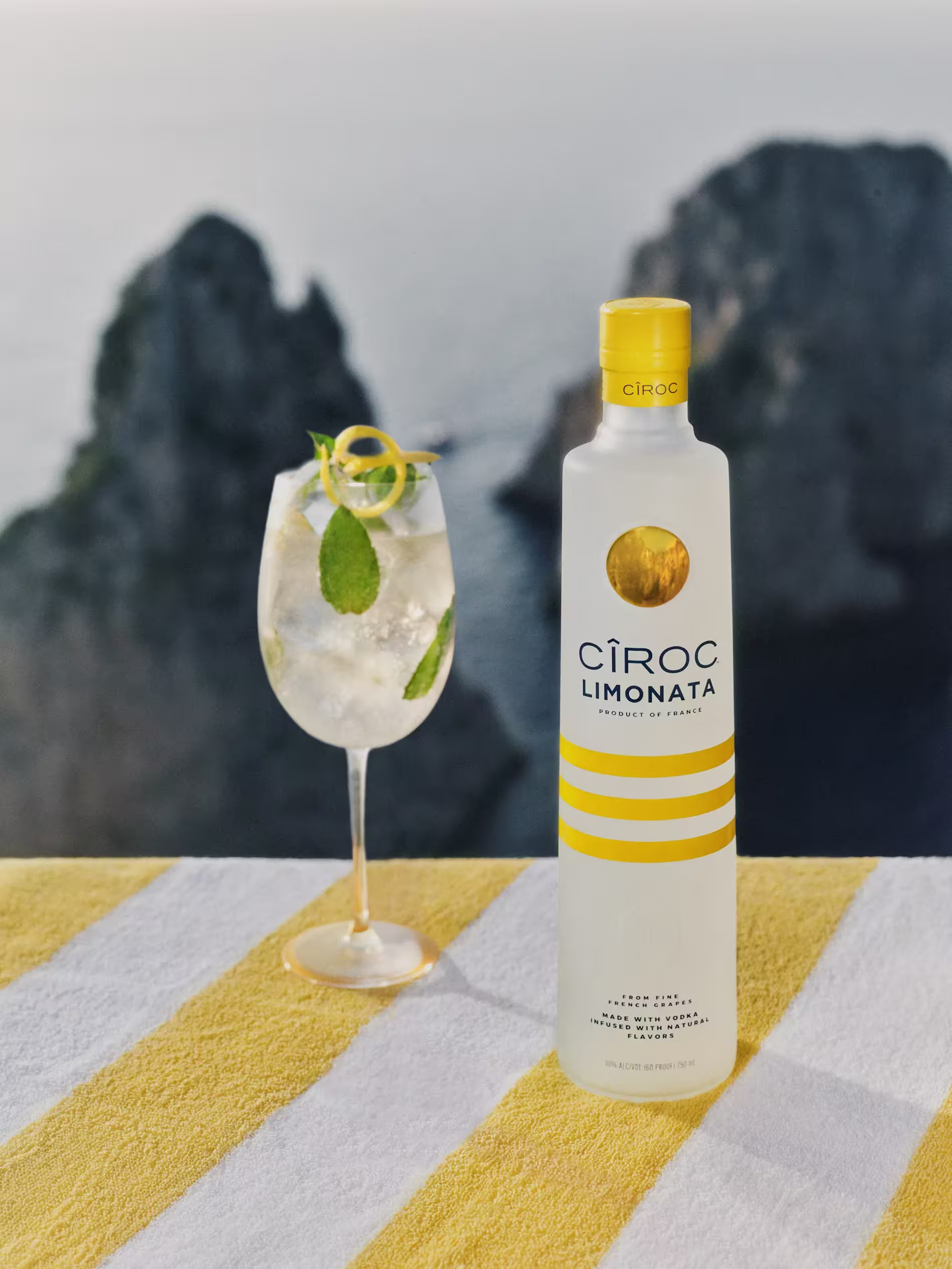 A bottle of Cîroc Limonata sits on a yellow and white striped towel next to a cocktail glass with ice, mint, and lemon, with rocky cliffs and water in the blurred background.