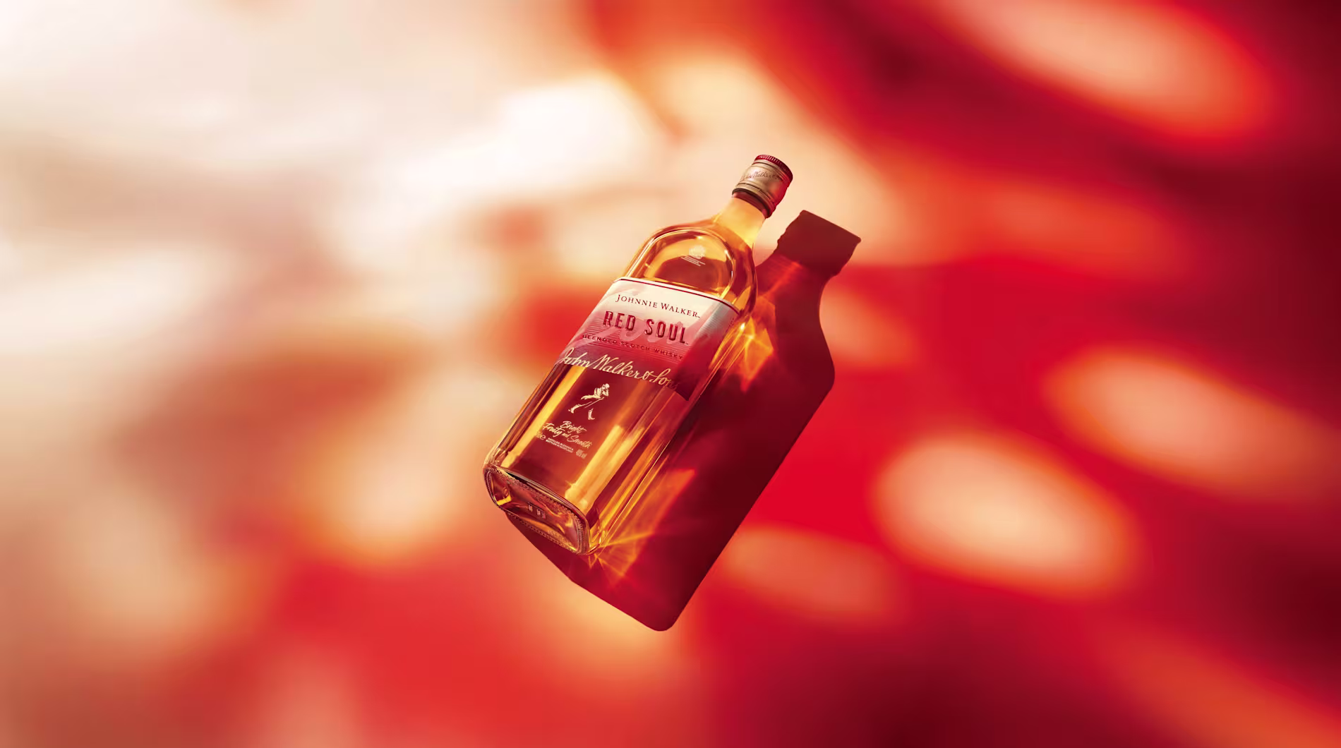 Una botella de whisky Johnnie Walker Red Sour, inclinada y iluminada con cálidos tonos ámbar, que flota sobre un fondo abstracto de vivos tonos rojos y naranjas, proyectando una sombra nítida hacia la derecha.
