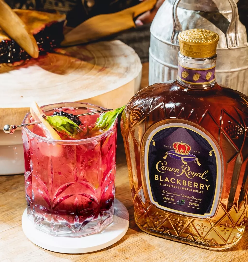 A glass of blackberry cocktail with garnish sits next to a bottle of Crown Royal Blackberry whisky on a wooden surface.