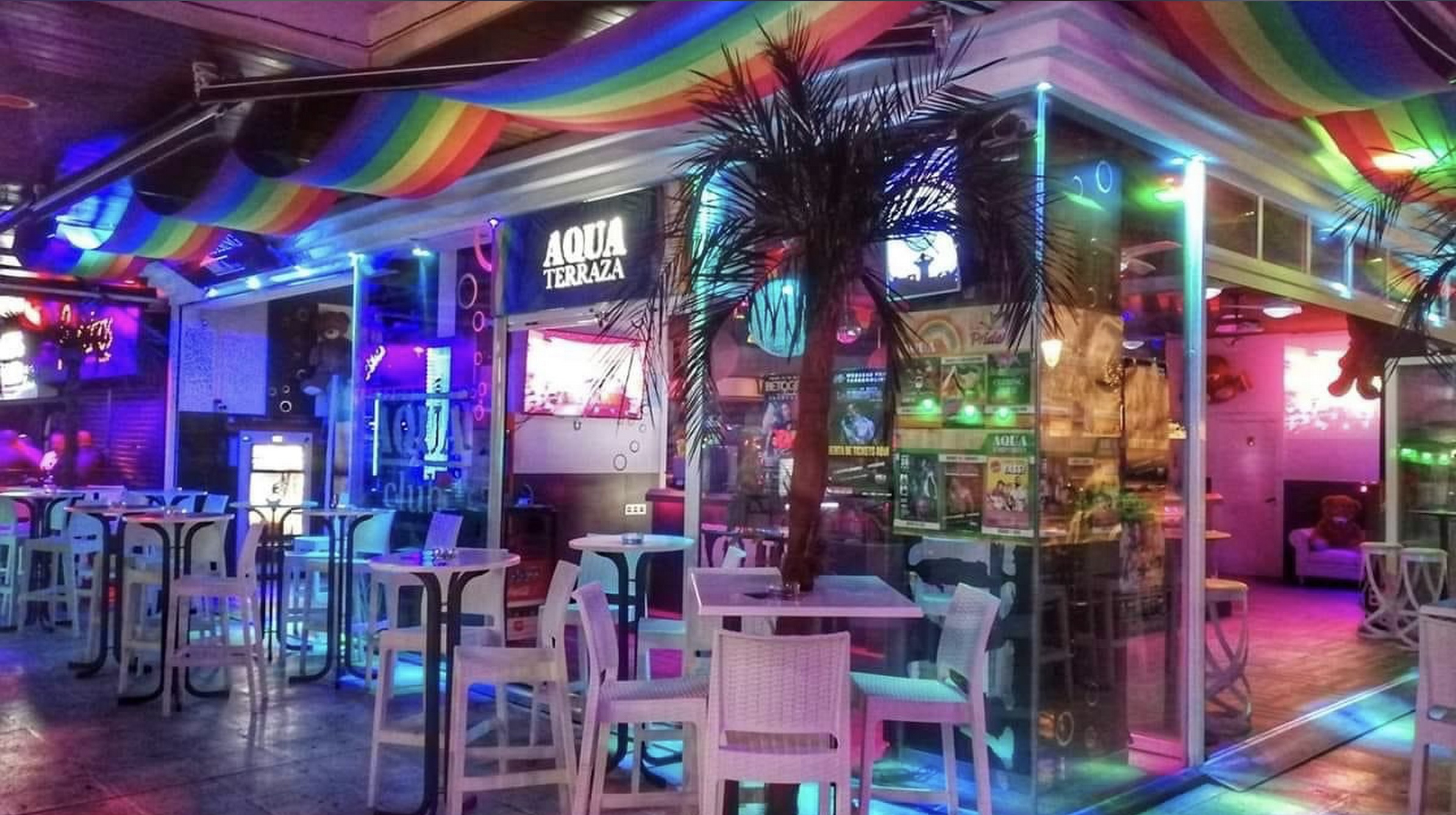 Terraza de Aqua Club Torremolinos, decorada con luces de neón, mesas blancas, palmeras y una bandera arcoíris en el techo.
