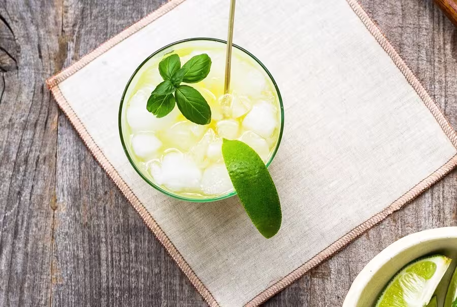 A glass of iced yellow drink garnished with a lime wedge and basil leaves sits on a cloth napkin atop a wooden surface.