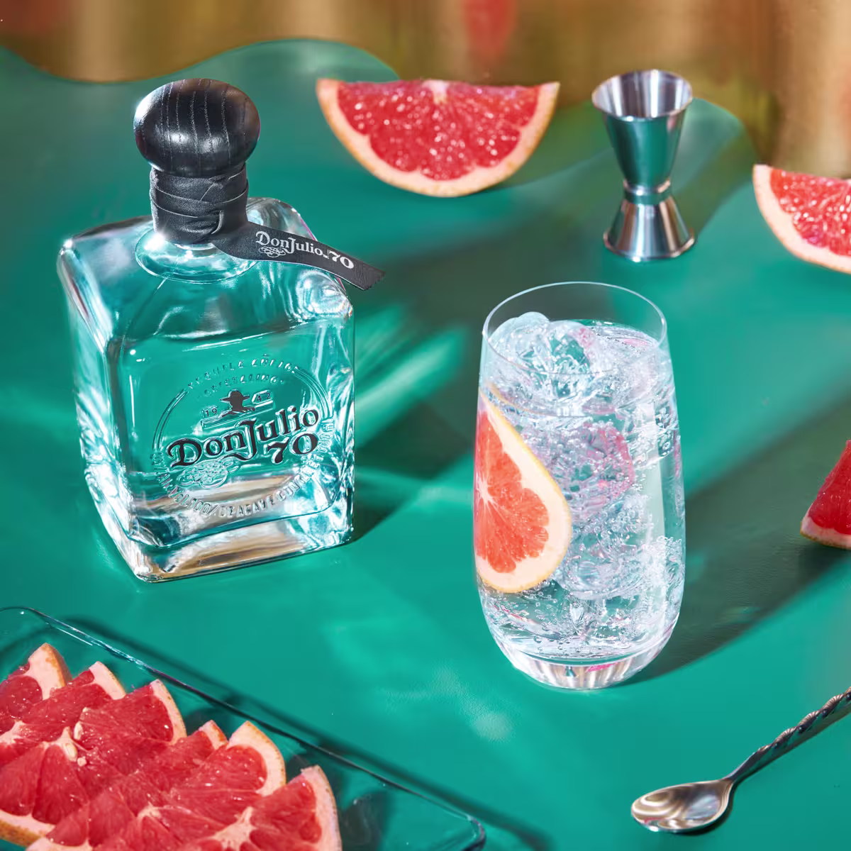A bottle of Don Julio 70 tequila, a glass with ice and grapefruit slices, grapefruit wedges, a jigger, and a spoon arranged on a green surface.