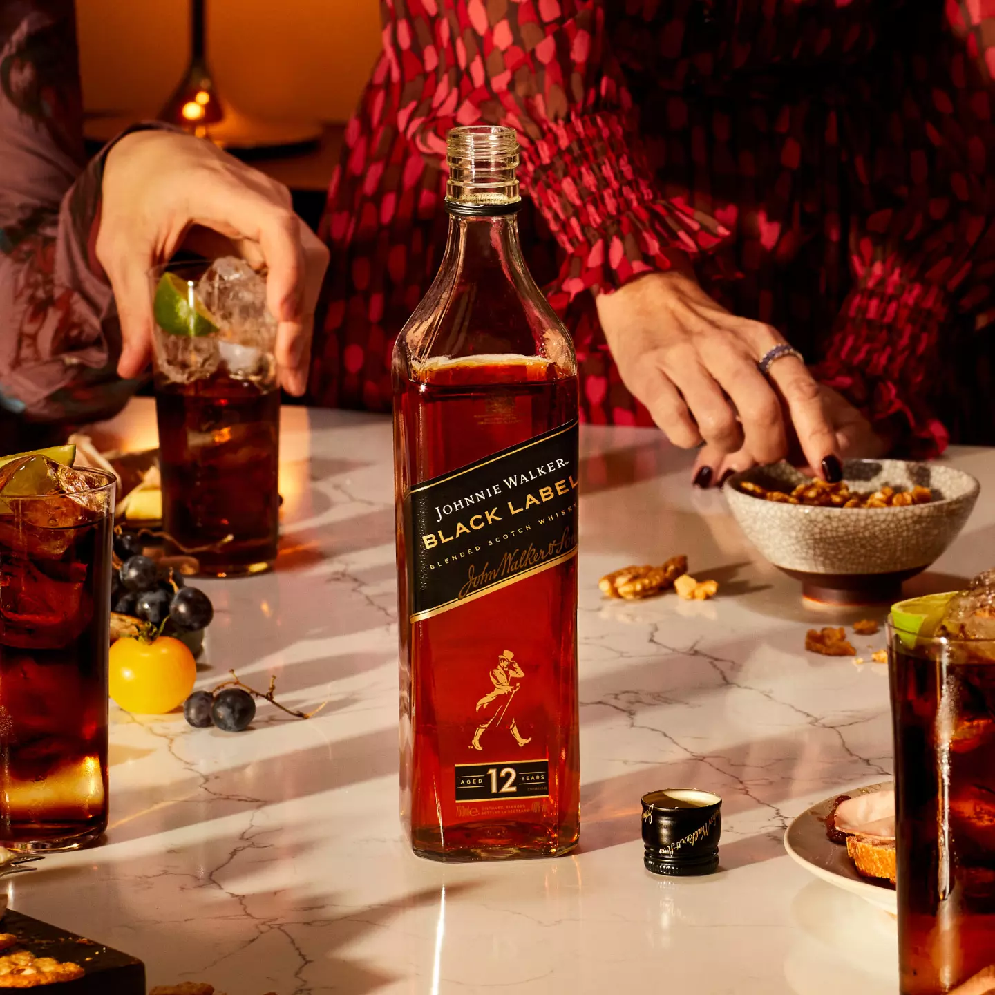 A bottle of Johnnie Walker Black Label whisky on a marble table surrounded by glasses of mixed drinks, snacks, and hands reaching for food.