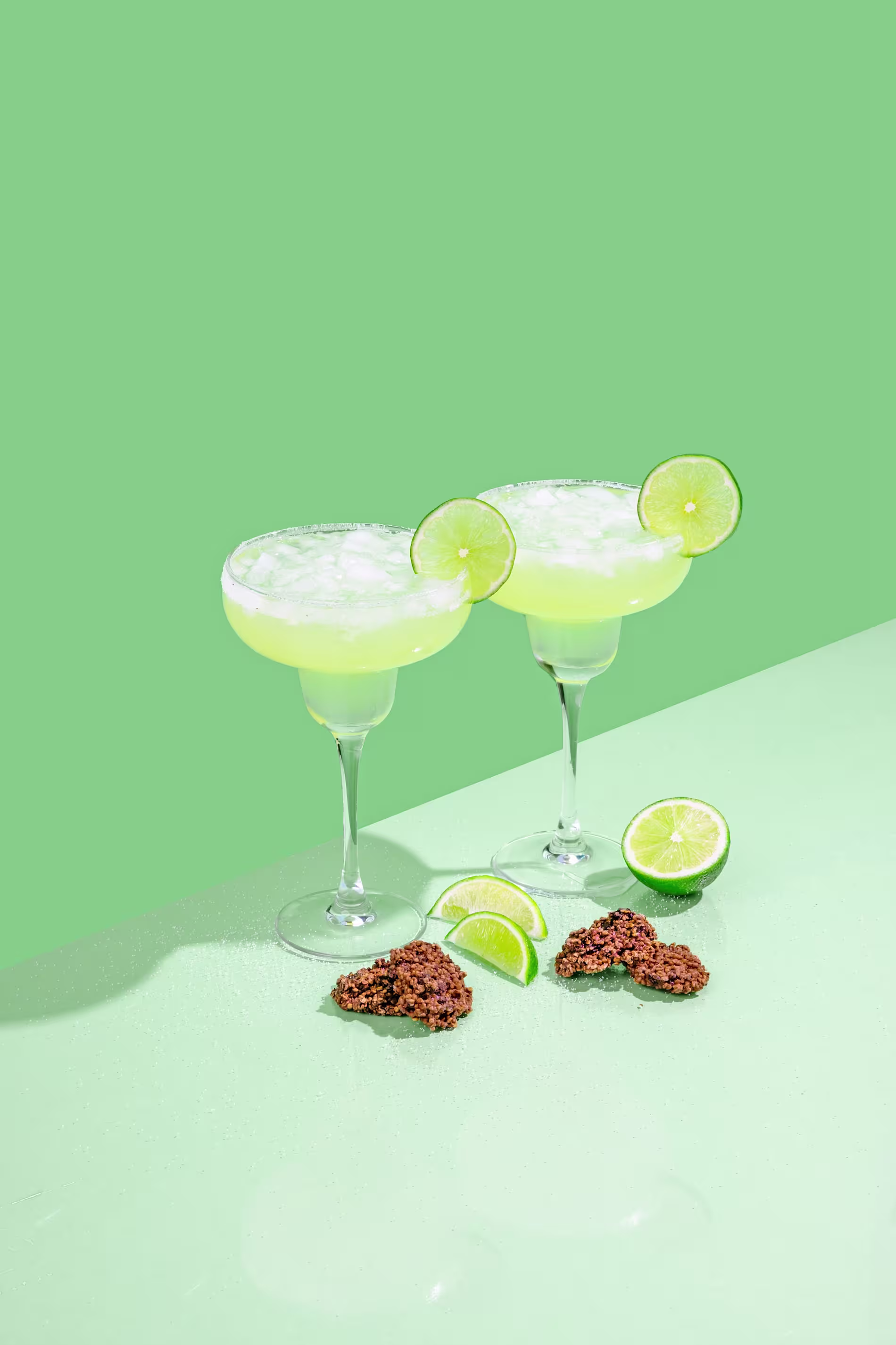Two margarita glasses with salted rims and lime slices, surrounded by lime wedges and chocolate clusters on a green surface with a green background.