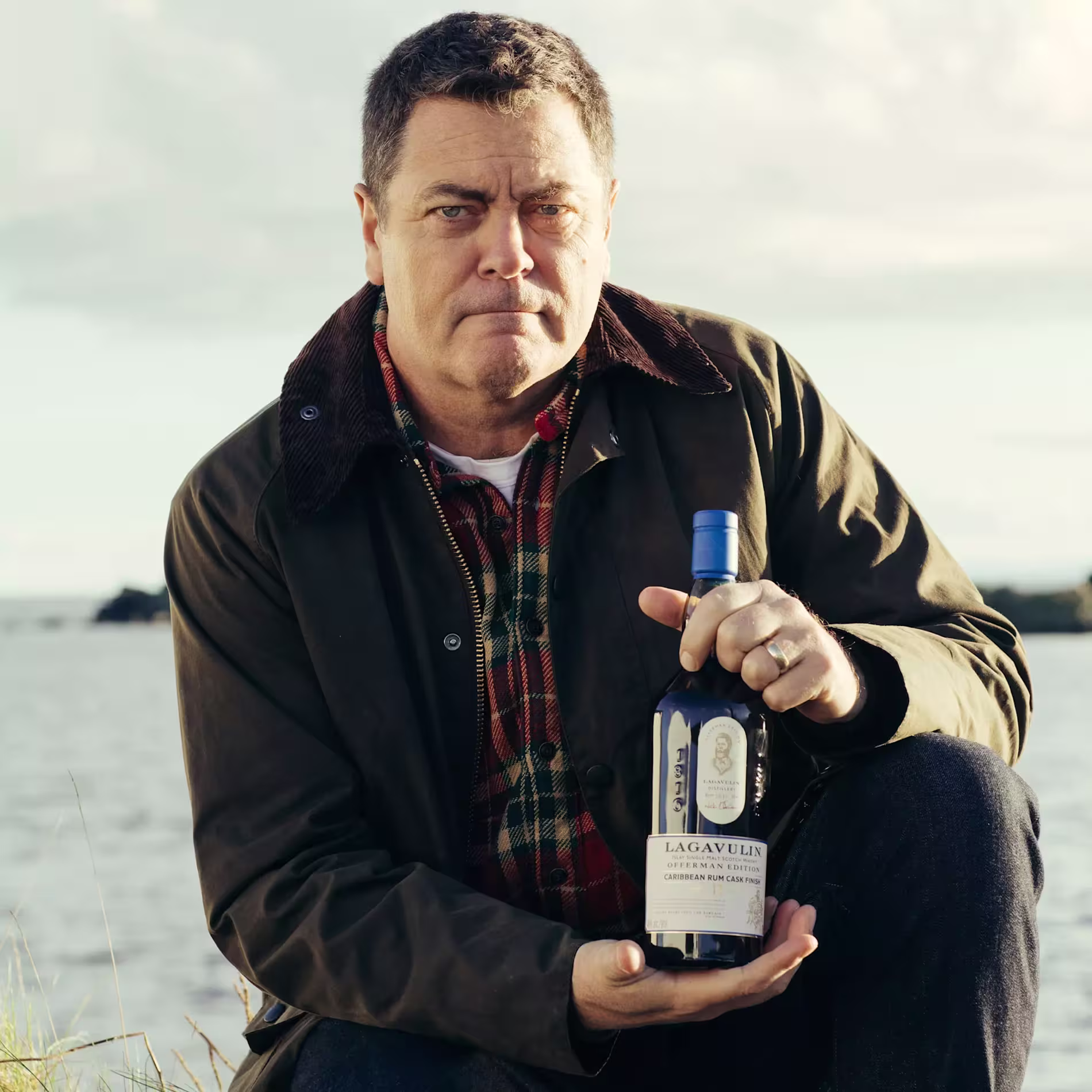 A man in an outdoor setting holds a bottle of Lagavulin whisky, wearing a green jacket over a plaid shirt.