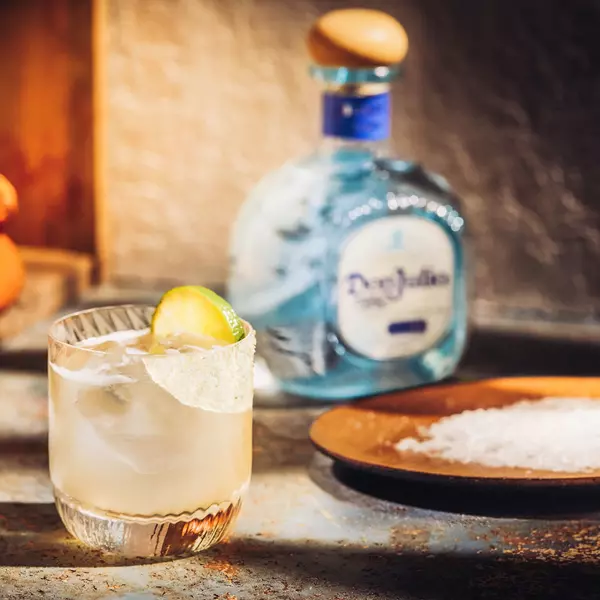 A glass of cocktail with ice, a lime wedge, and a salted rim sits on a table near a plate of salt and a bottle of Don Julio tequila in the background.