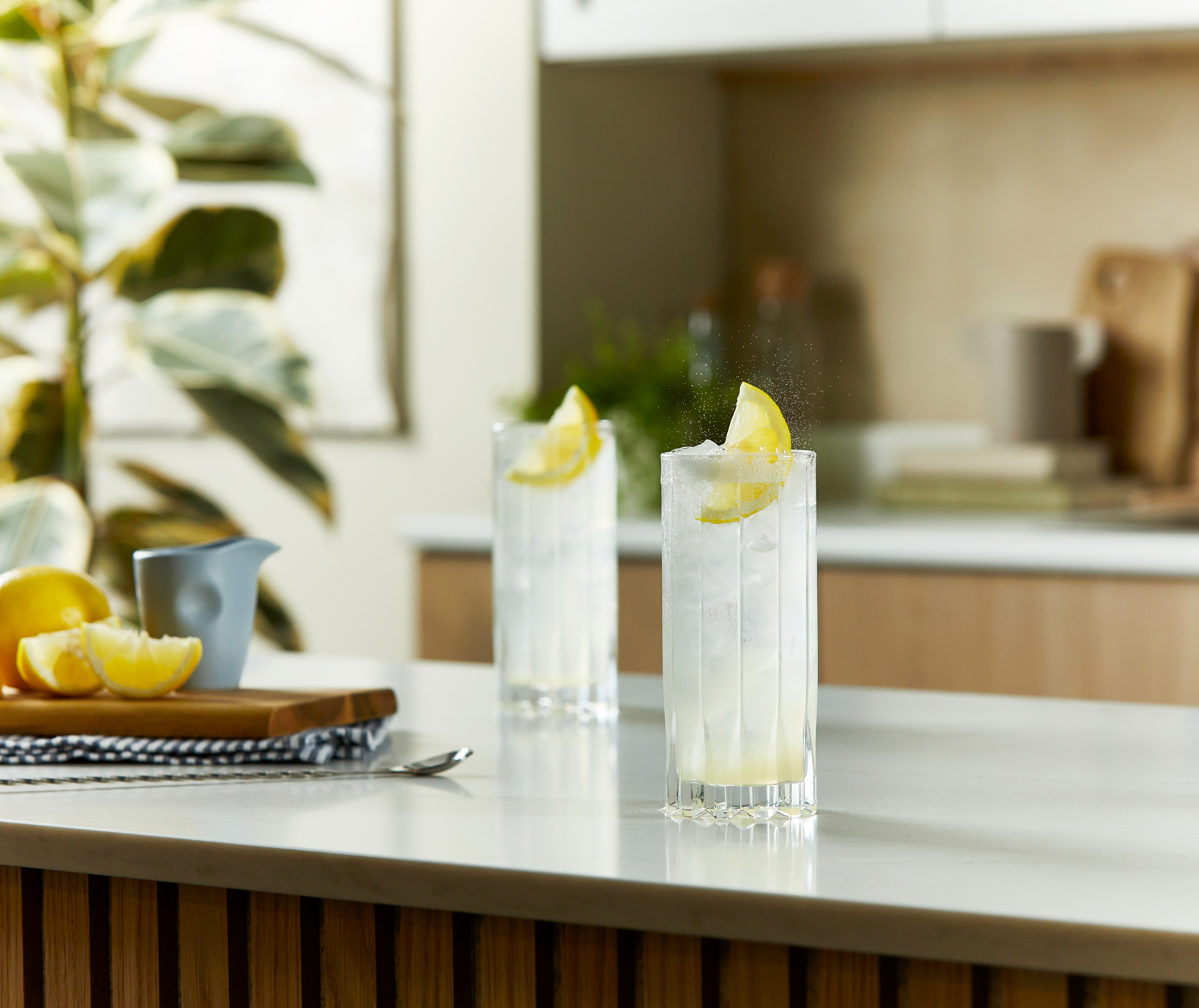 Two Tom Collins cocktails sit on a kitchen counter, in highball glasses with lots of ice and lemon wedges.