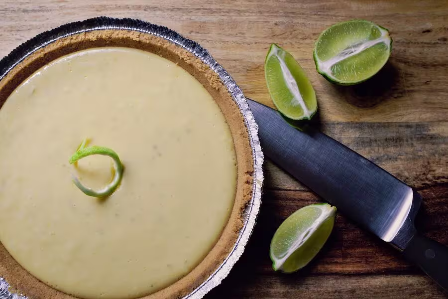 A key lime pie in a foil pan sits on a wooden surface next to a knife and two halved limes.