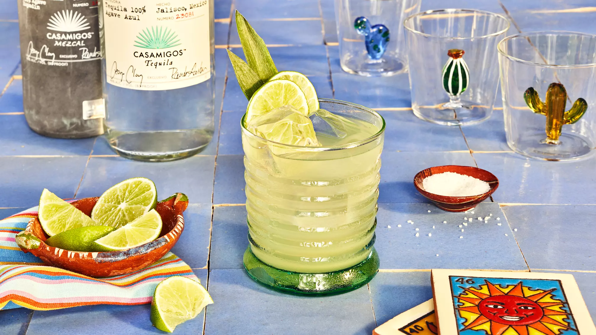 A margarita garnished with lime and salt sits on a blue tiled table, surrounded by tequila bottles, lime wedges, salt, decorated glasses, and a lotería card.