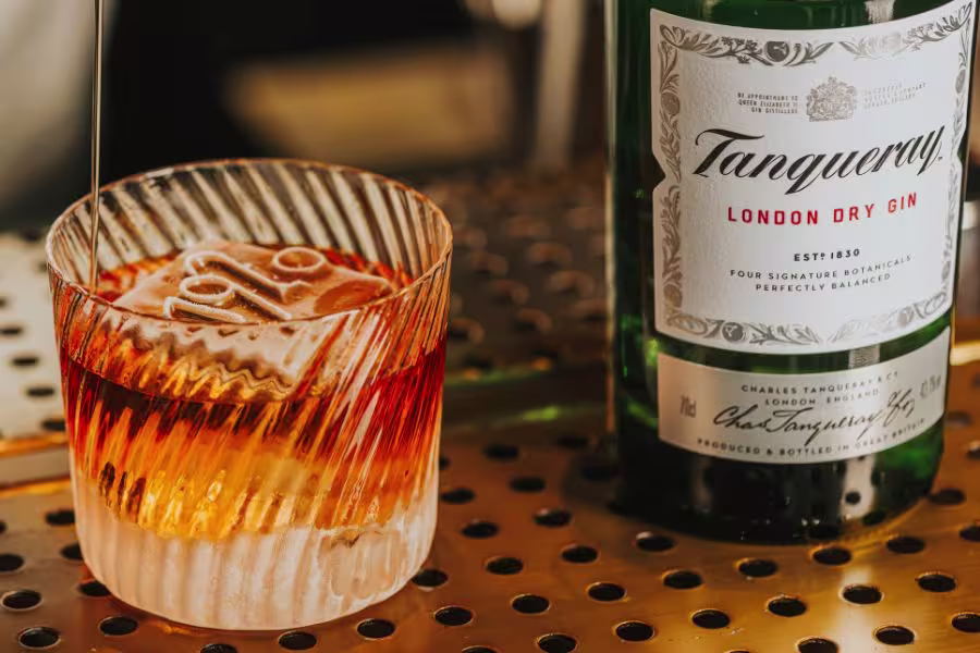 A glass of clear cocktail with ice, lime slices, berries, and a black straw sits on a wooden table in an outdoor setting.
