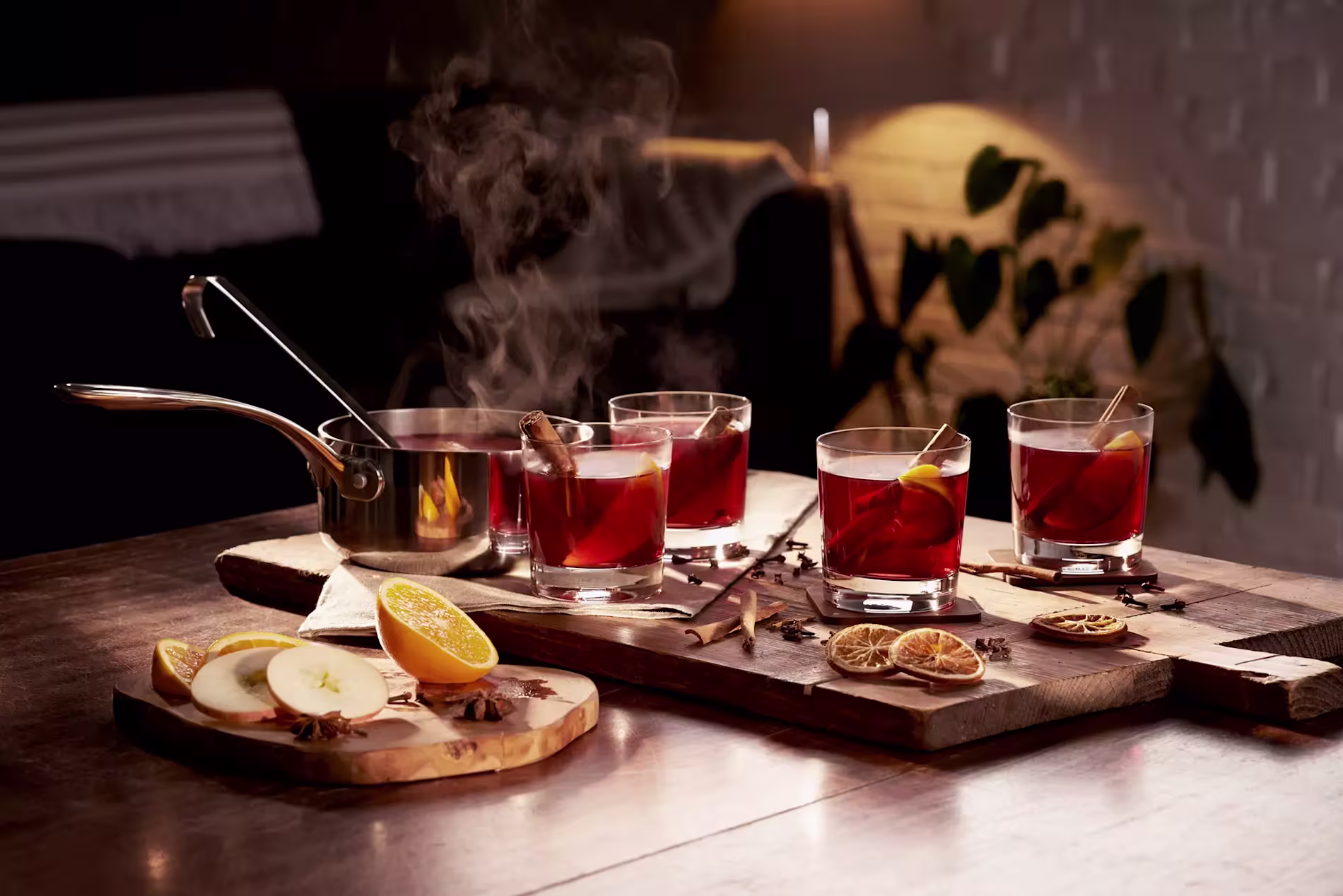 Four glasses of mulled wine with citrus and cinnamon are arranged on wooden boards, beside a steaming saucepan and slices of apple, orange, and spices on a table.