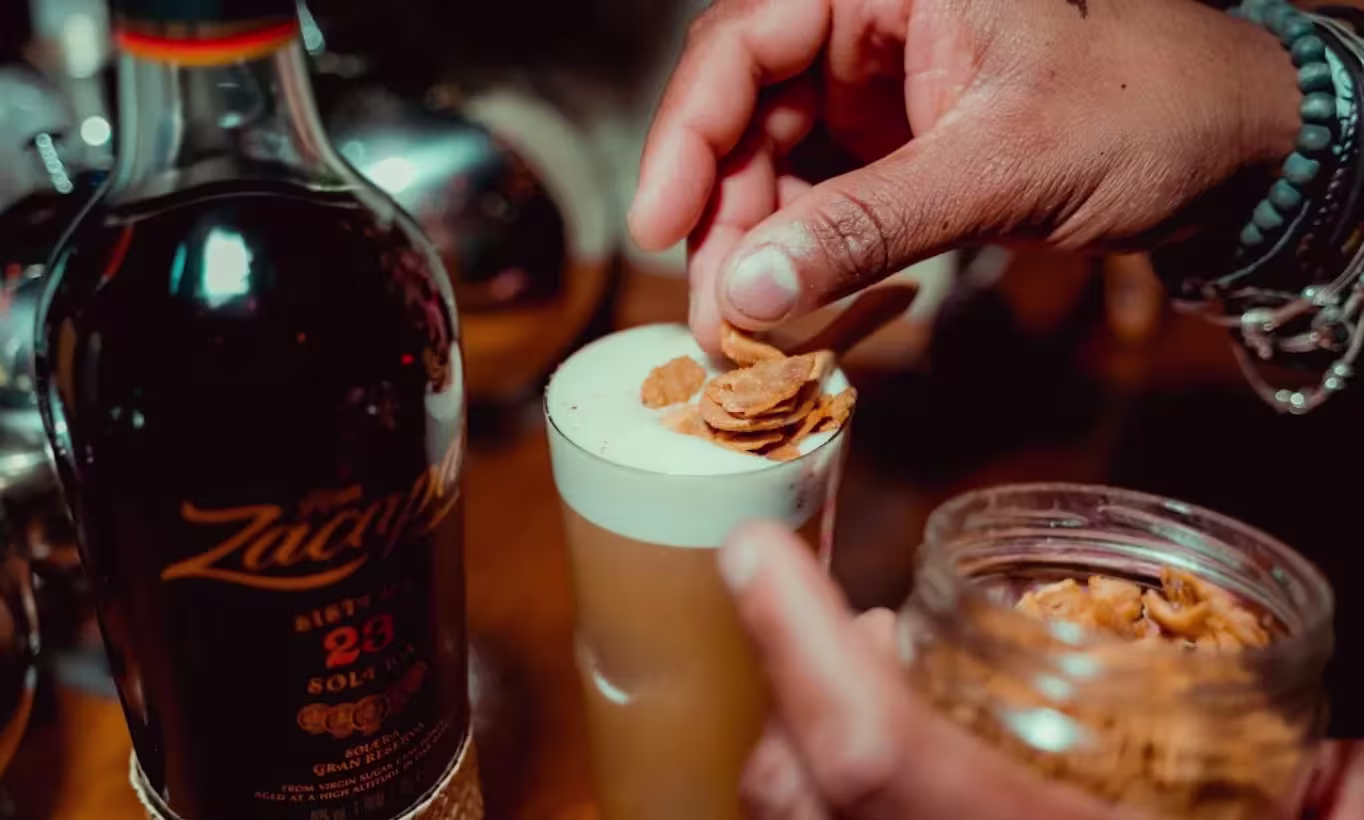La imagen muestra la preparación de un cóctel con Ron Zacapa, decorado con cereales, mientras se añaden con una mano al vaso.