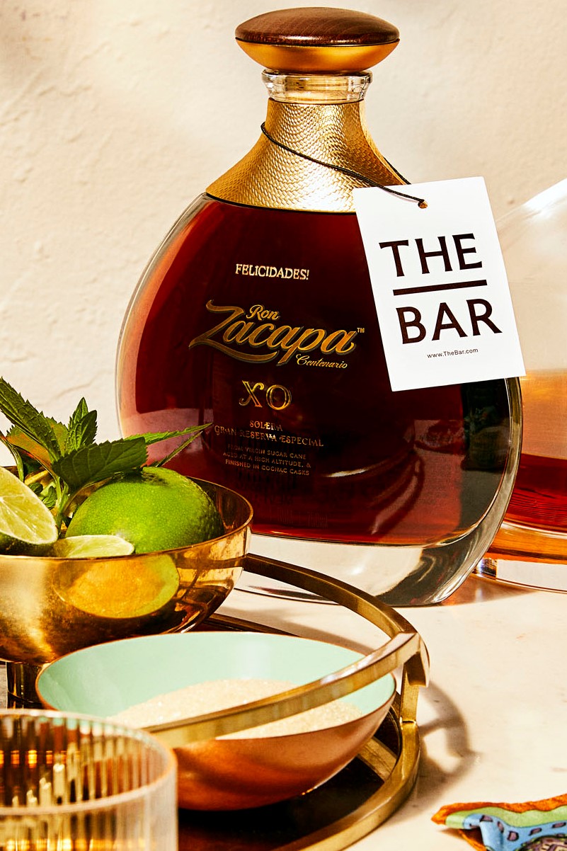 A bottle of Ron Zacapa XO rum with a tag labeled The Bar stands on a tray next to a bowl of mint, limes, and a turquoise dish.