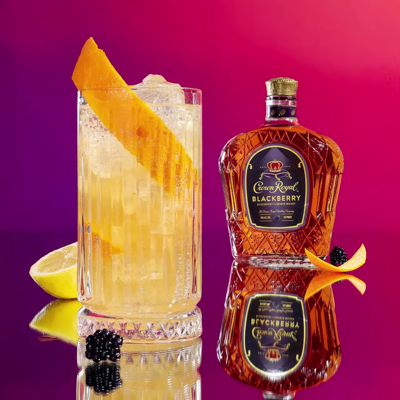 A glass of cocktail garnished with an orange peel sits next to a bottle of Crown Royal Blackberry whisky, with a blackberry and orange peel placed nearby against a pink gradient background.