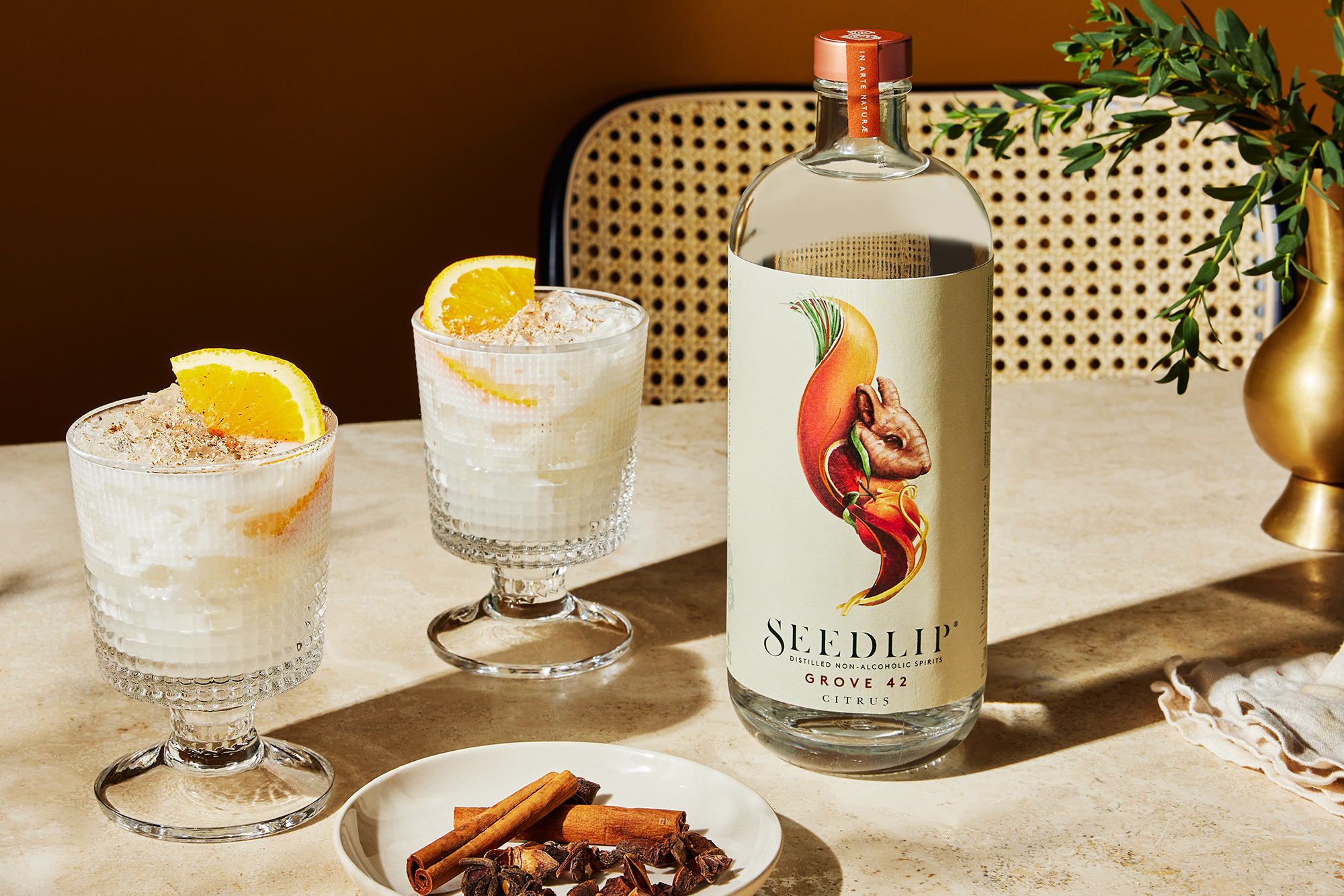 A bottle of Seedlip Grove 42 non-alcoholic spirit is on a table beside two creamy cocktails garnished with orange slices, with a small plate of cinnamon sticks and star anise in front.