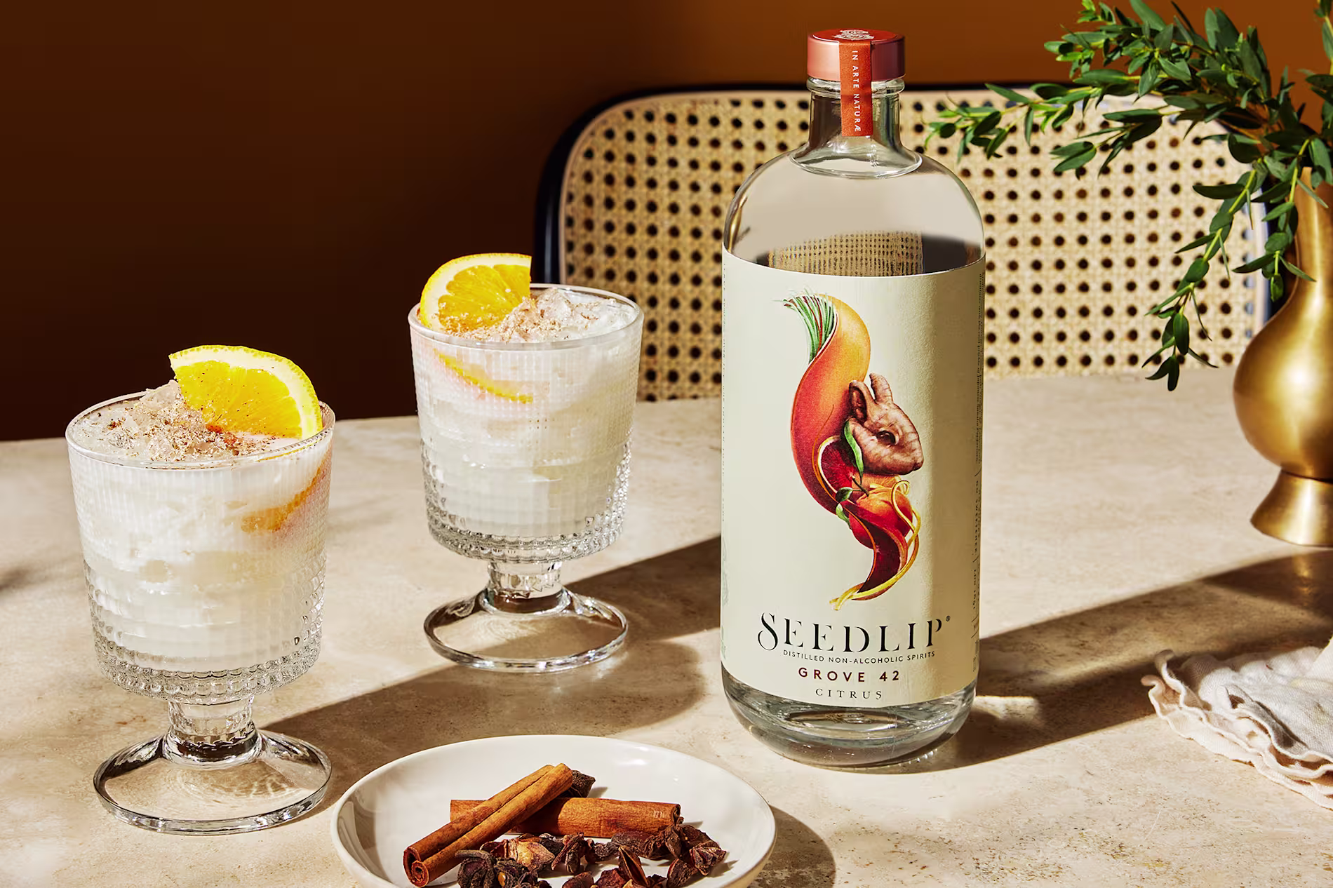 A bottle of Seedlip Grove 42 non-alcoholic spirit is on a table beside two creamy cocktails garnished with orange slices, with a small plate of cinnamon sticks and star anise in front.
