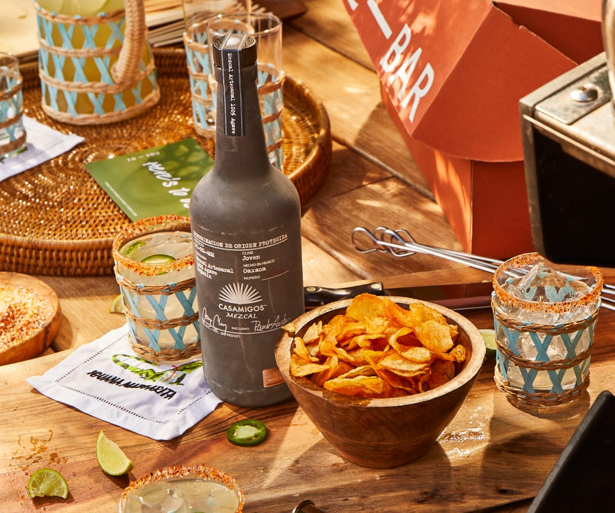 A bottle of Casamigos mezcal, a wooden bowl of chips, and cocktails on a sunlit wooden table set for outdoor entertaining.