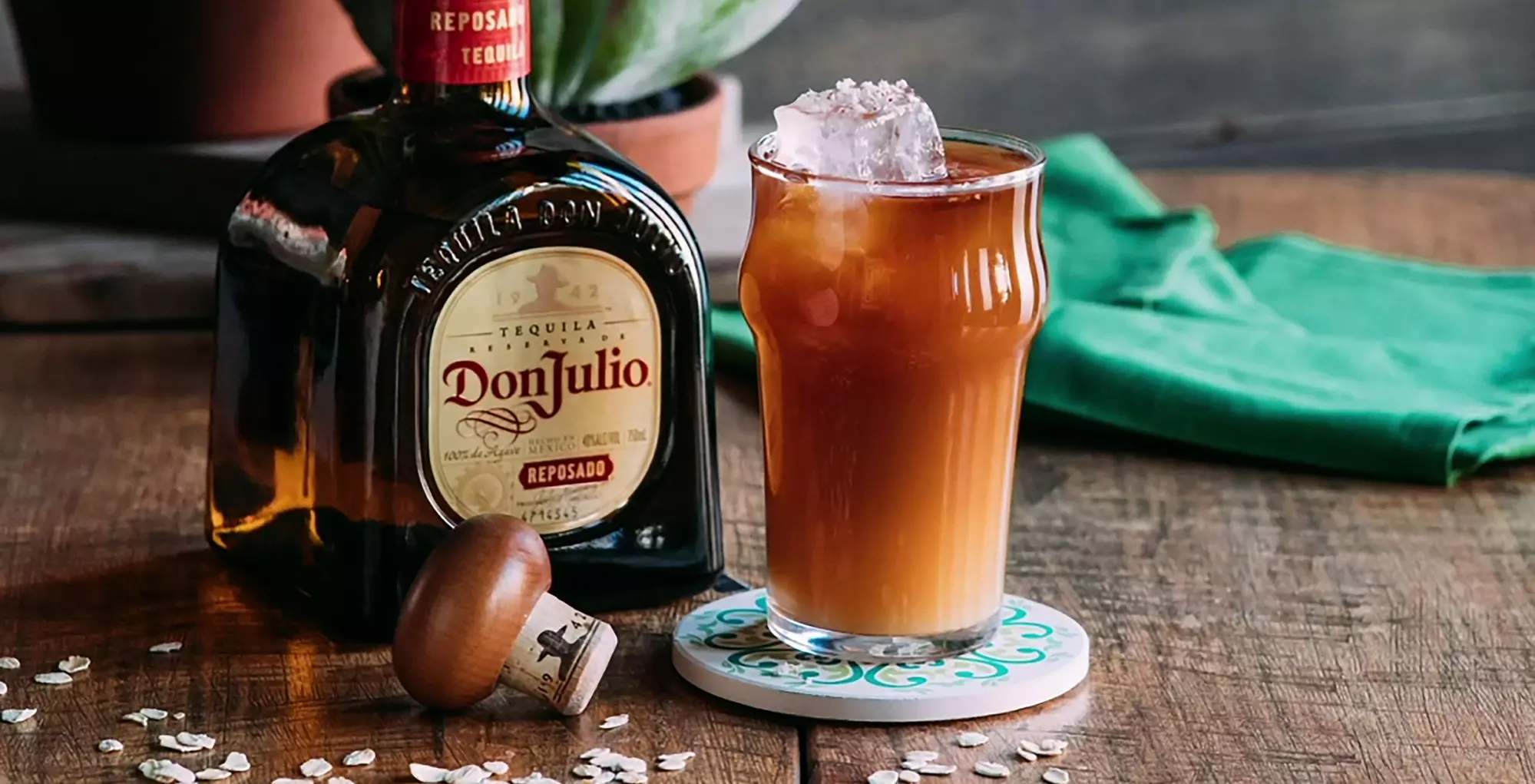 A bottle of Don Julio Reposado tequila stands next to a glass of iced brown cocktail on a coaster, with a cork and some oats on a wooden surface.
