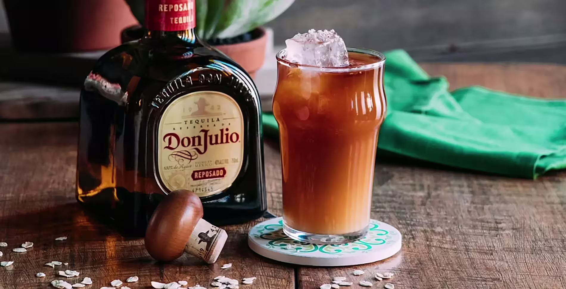 A bottle of Don Julio Reposado tequila stands next to a glass of iced brown cocktail on a coaster, with a cork and some oats on a wooden surface.