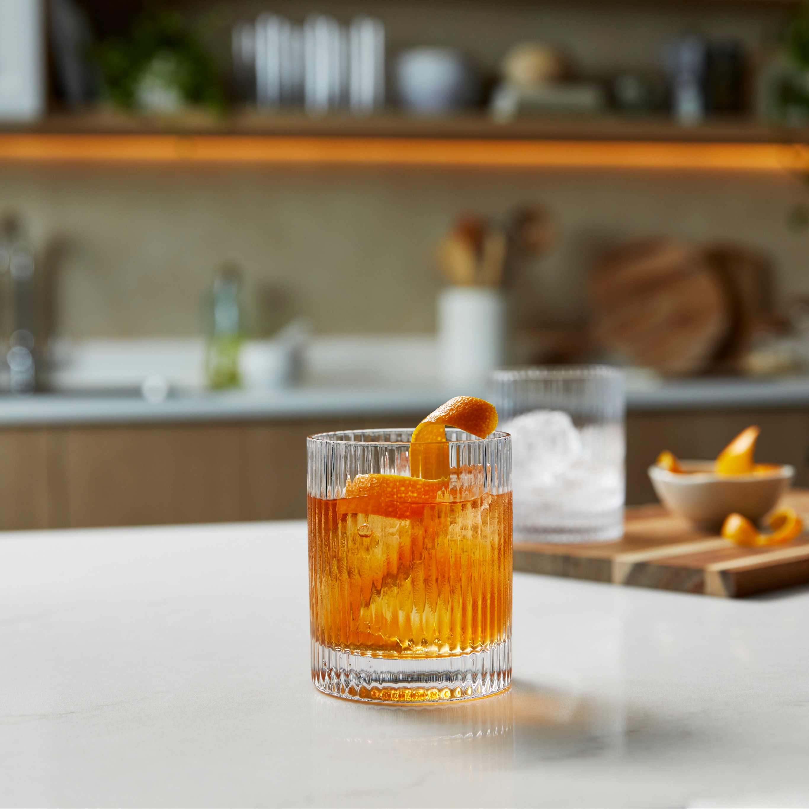 An Old Fashioned cocktails with a twisted orange garnish on a shiny white kitchen counter.